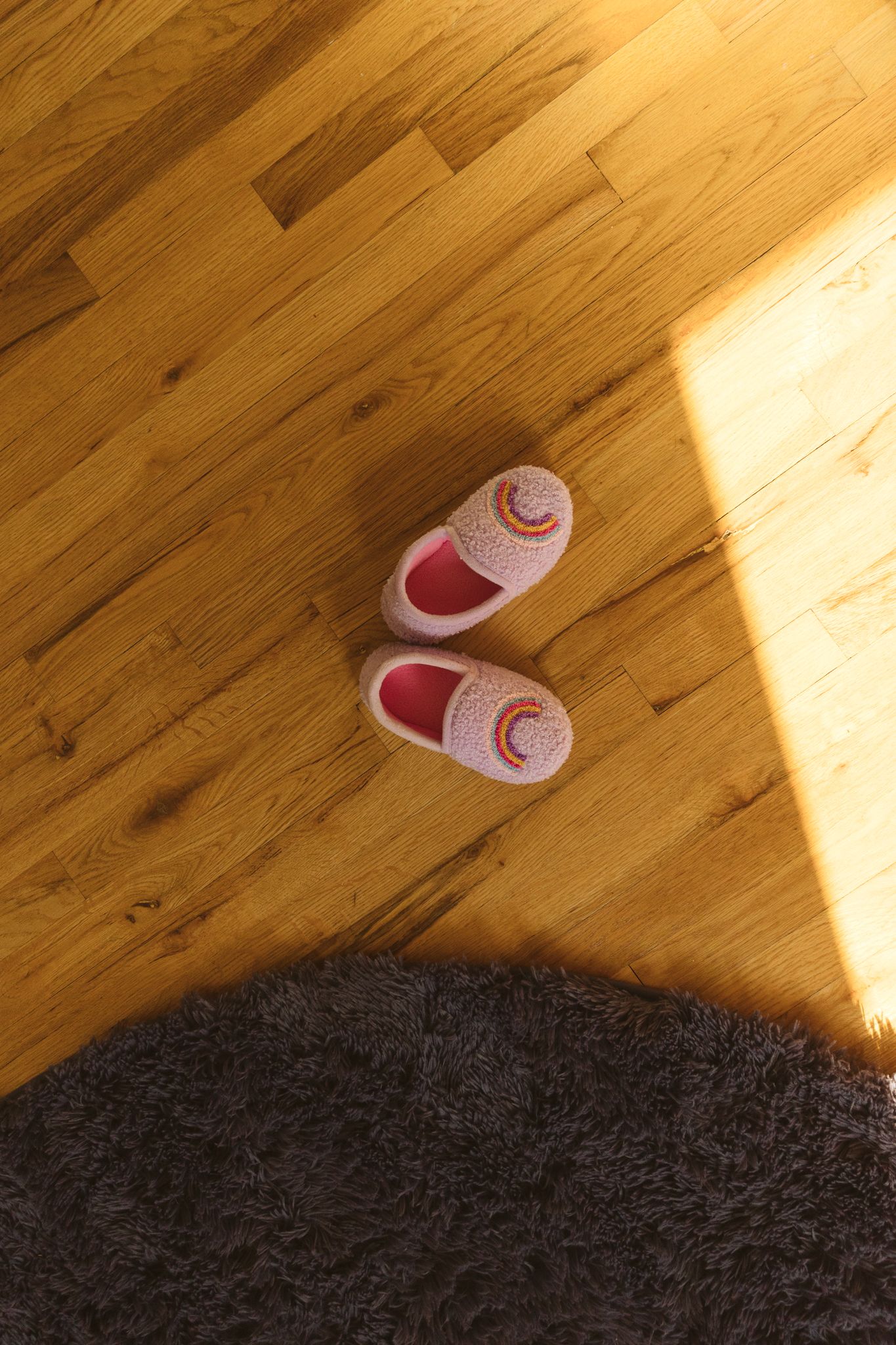 Pink rainbow slippers on warm wooden floor beside a gray fluffy rug in soft sunlight.