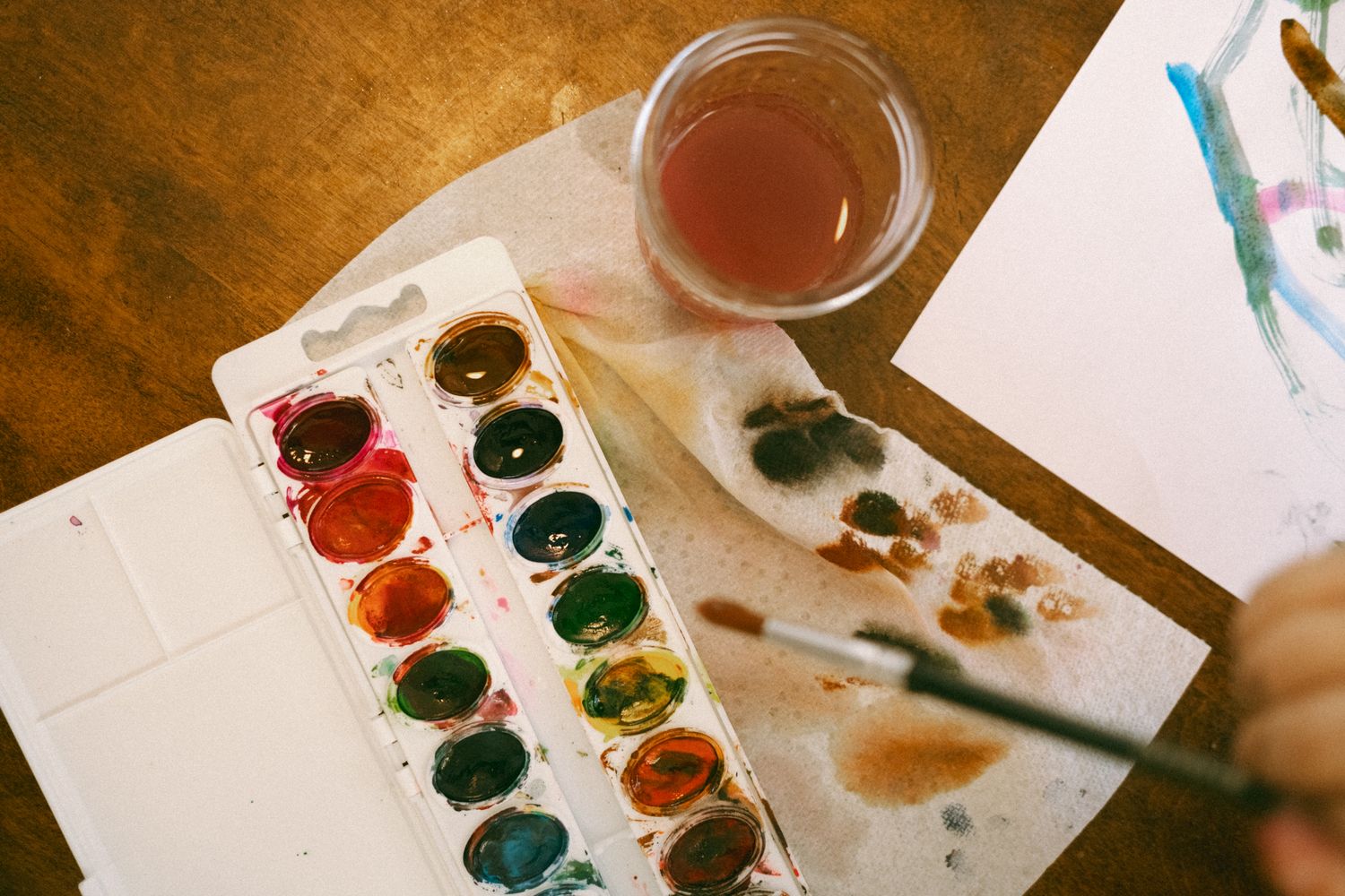Watercolor paint palette with vibrant colors, a brush, and a glass of water on a wooden table.