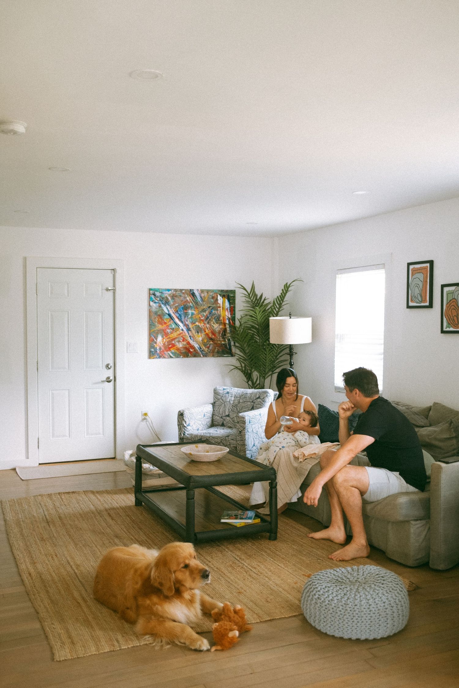 A couple relaxes in a bright living room with a golden retriever on a jute rug, colorful wall art, and modern furniture.