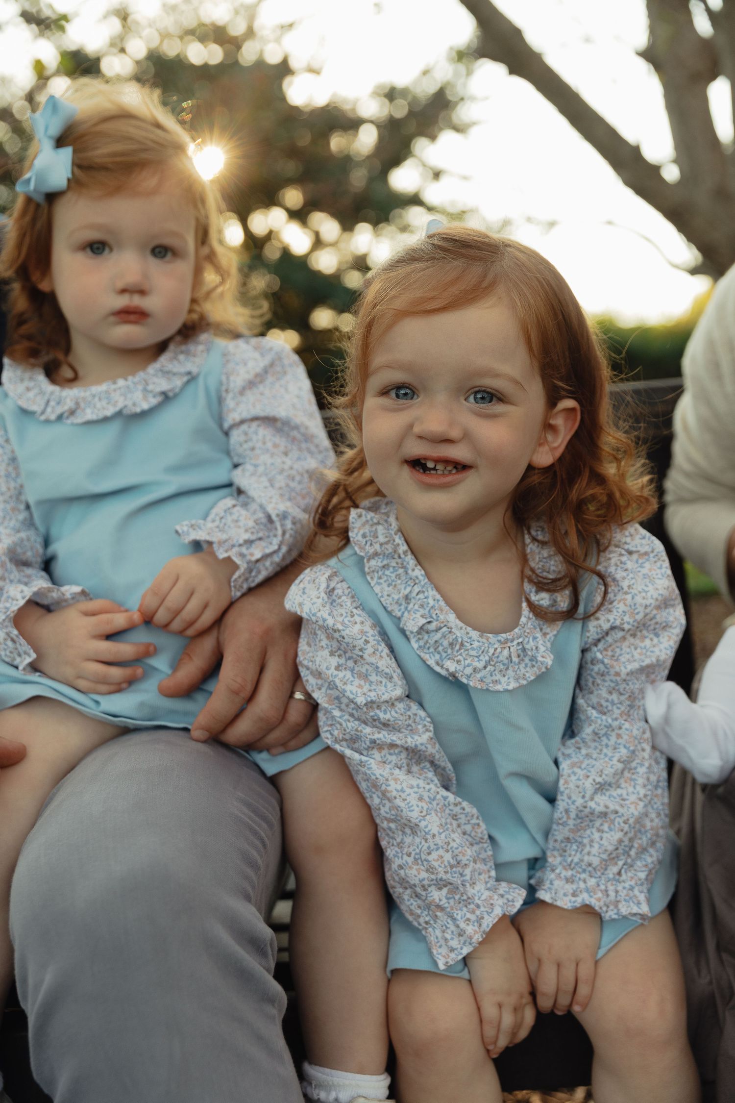 Two adorable redhead toddler sisters in matching blue dresses and floral tops outdoors.