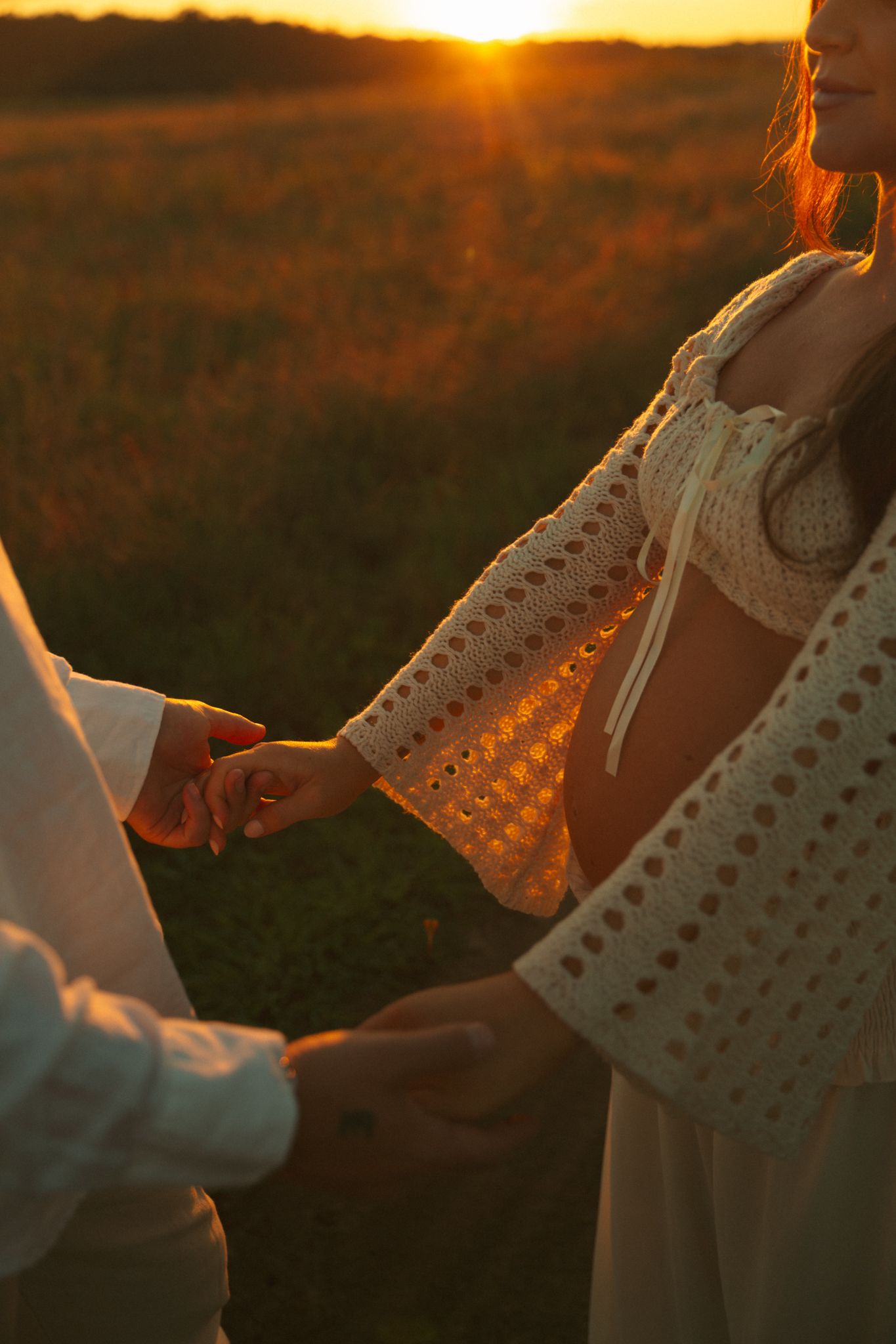 Couple holding hands in golden field during stunning sunset, woman wearing white crochet dress.