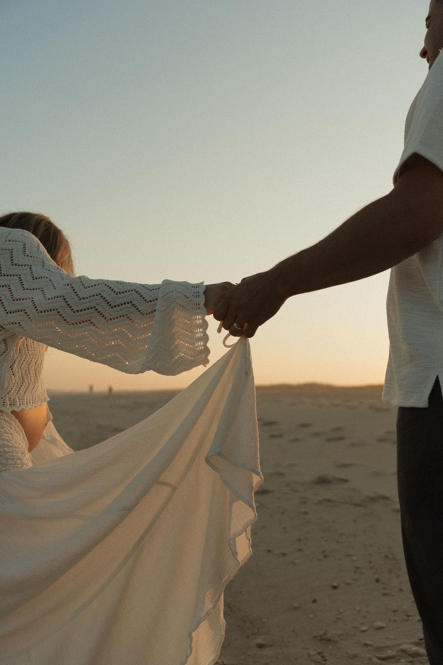 Couple holding hands on sandy beach at sunset, woman in white lace dress with flowing fabric.