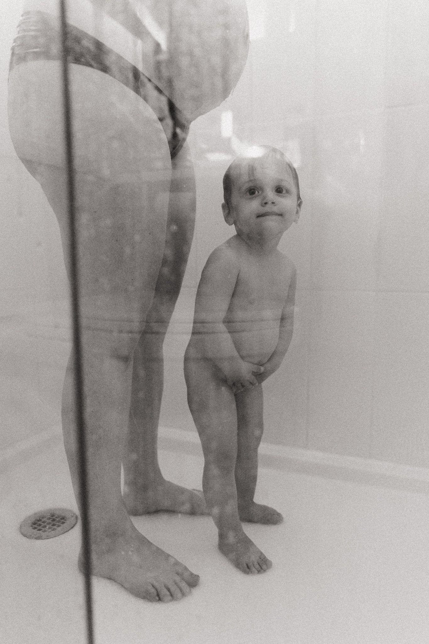 Black and white photo of a toddler standing in a shower with an adult, looking up curiously.
