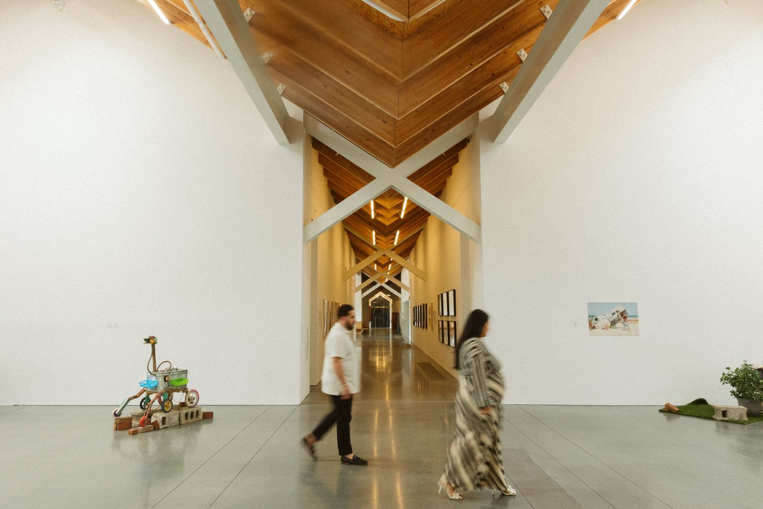 Modern art gallery interior with wooden chevron ceiling, white walls, artwork displays, and visitors walking through.