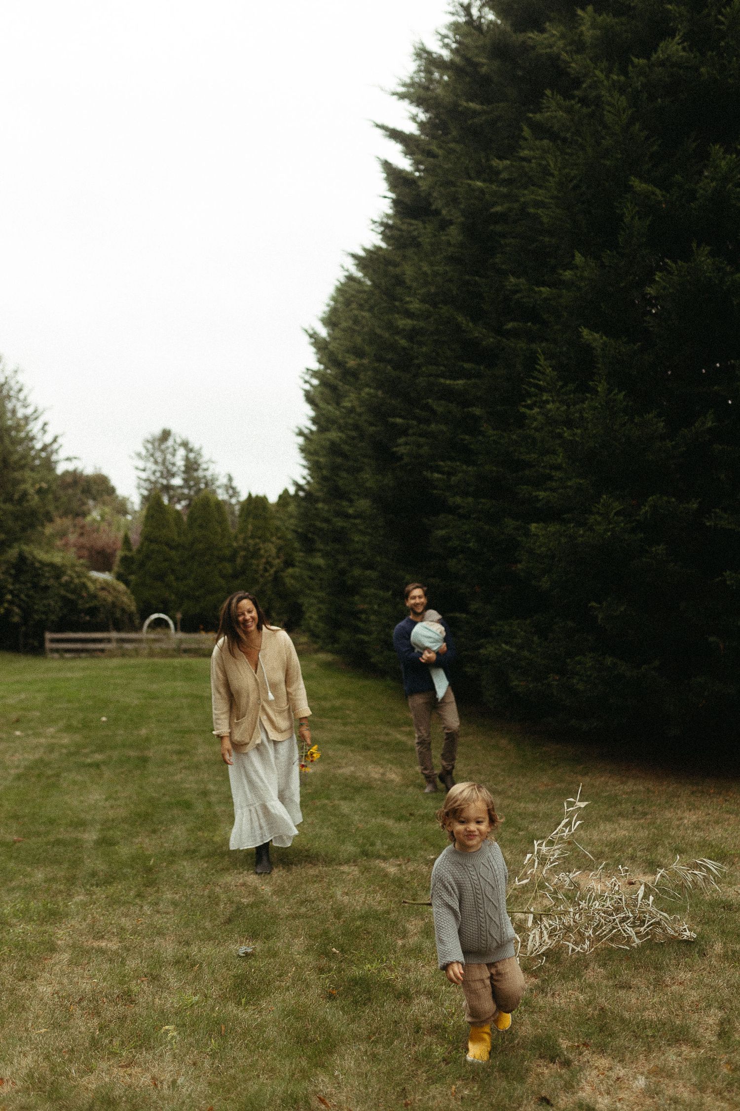 A family enjoys a casual stroll on a grassy lawn bordered by tall evergreen trees.