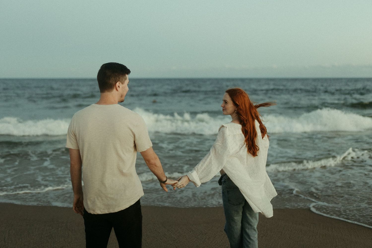 A romantic couple walks hand in hand along a misty beach with waves crashing behind them.