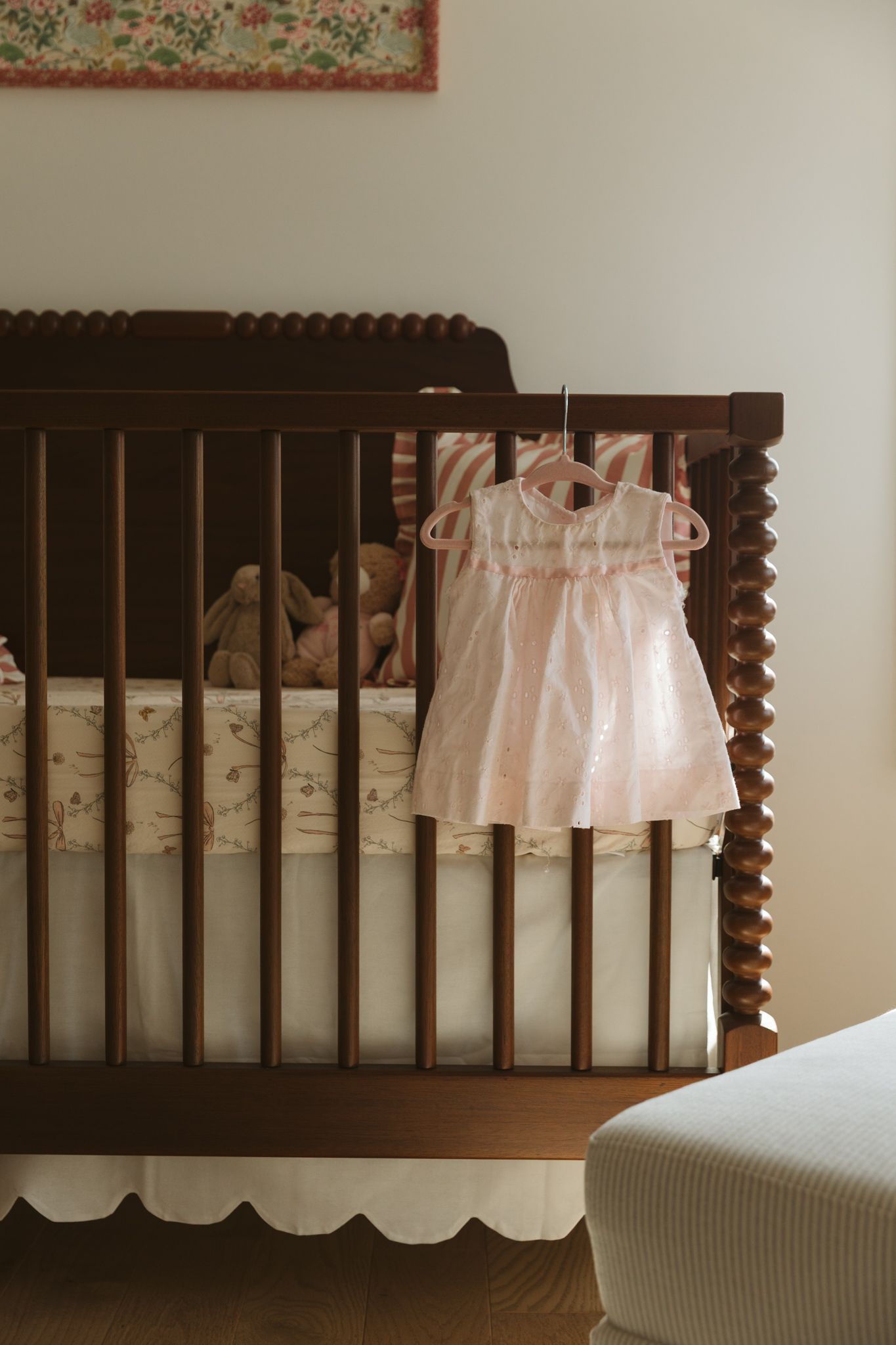 Dark wooden crib with white dress and floral artwork in a neutral nursery setting.