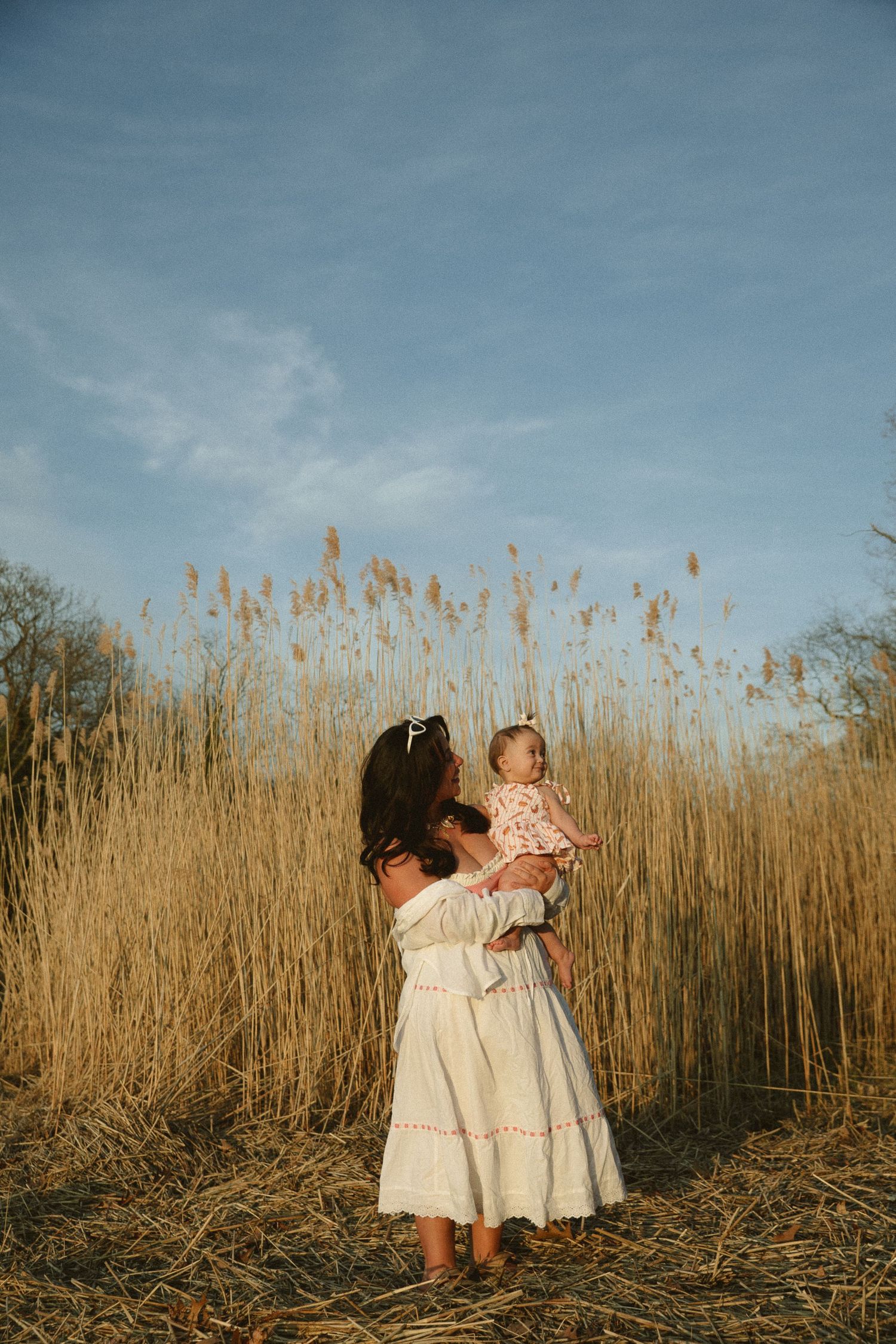 A mother in a white dress holds her baby in a golden reed field under a blue sky.