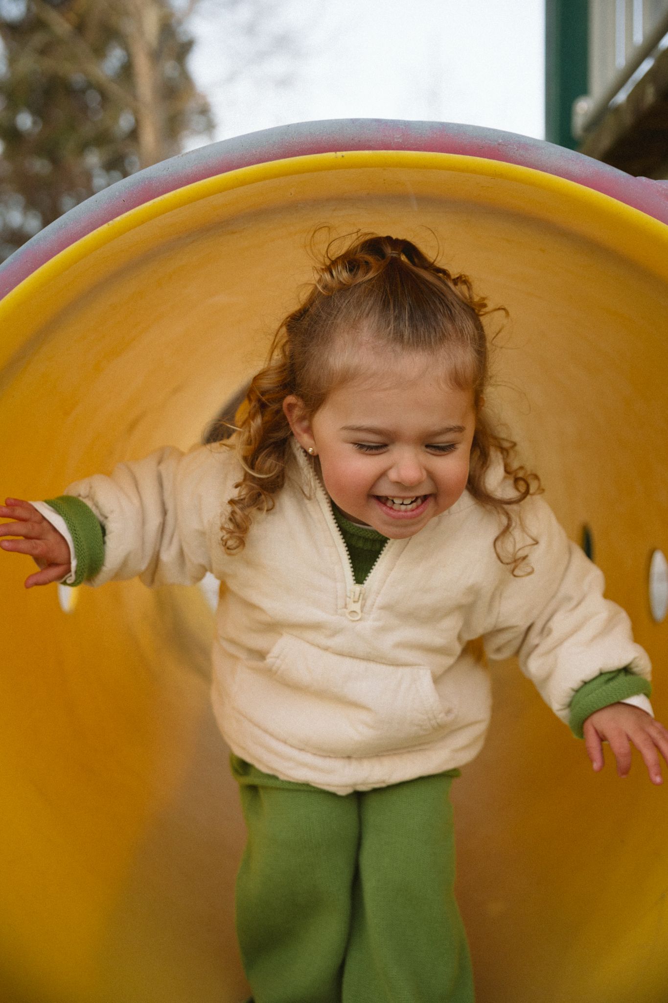 Joyful curly-haired toddler girl laughing while sliding through yellow tunnel on playground.