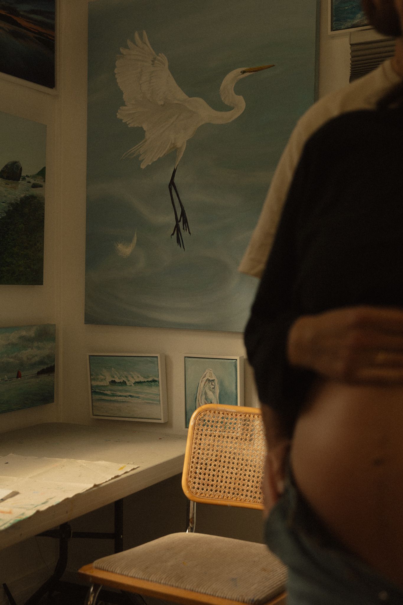 Pregnant woman viewing large egret painting in warmly lit art studio with coastal artwork display.