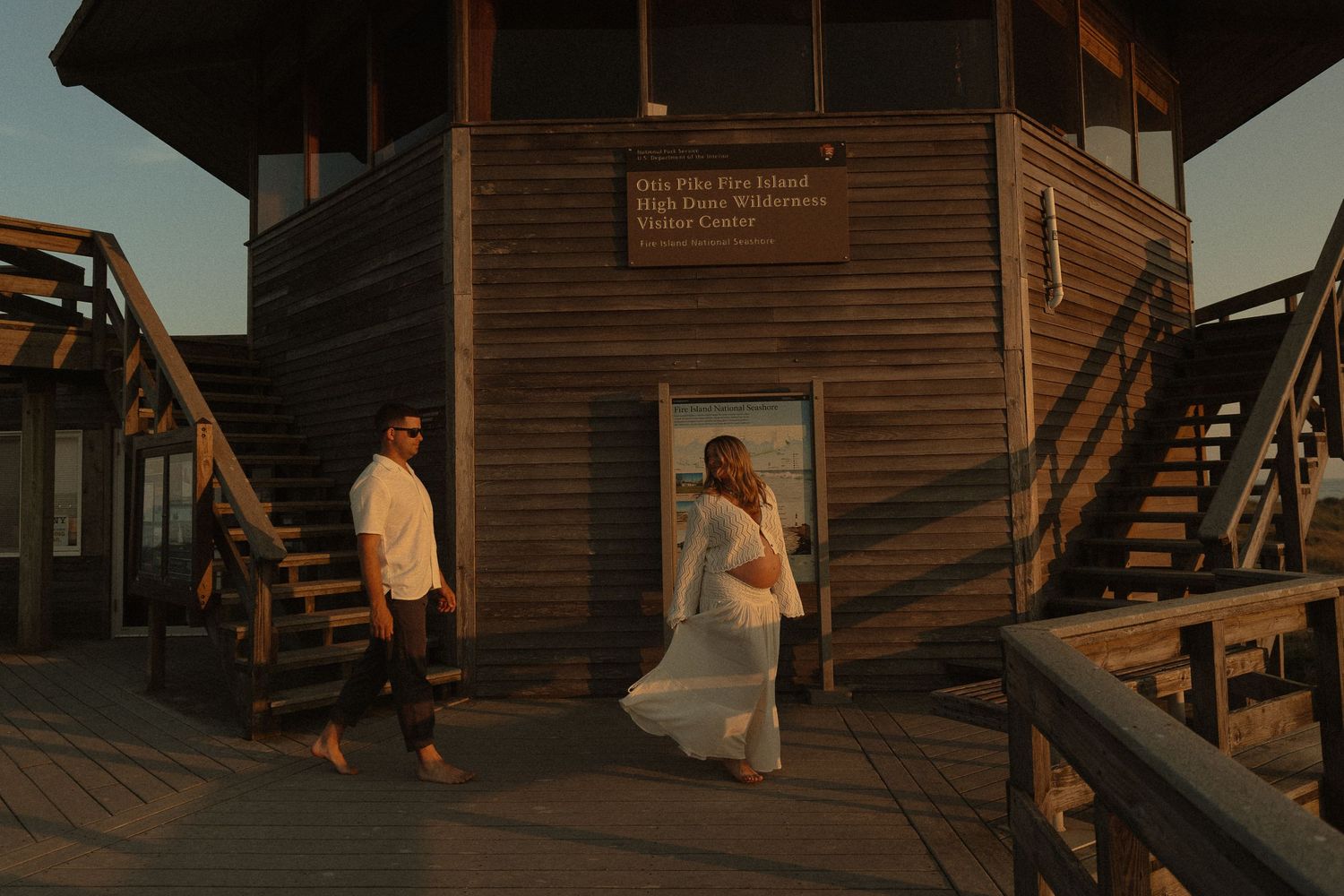 Expecting couple walking near a wooden building at Fire Island at golden hour sunset.