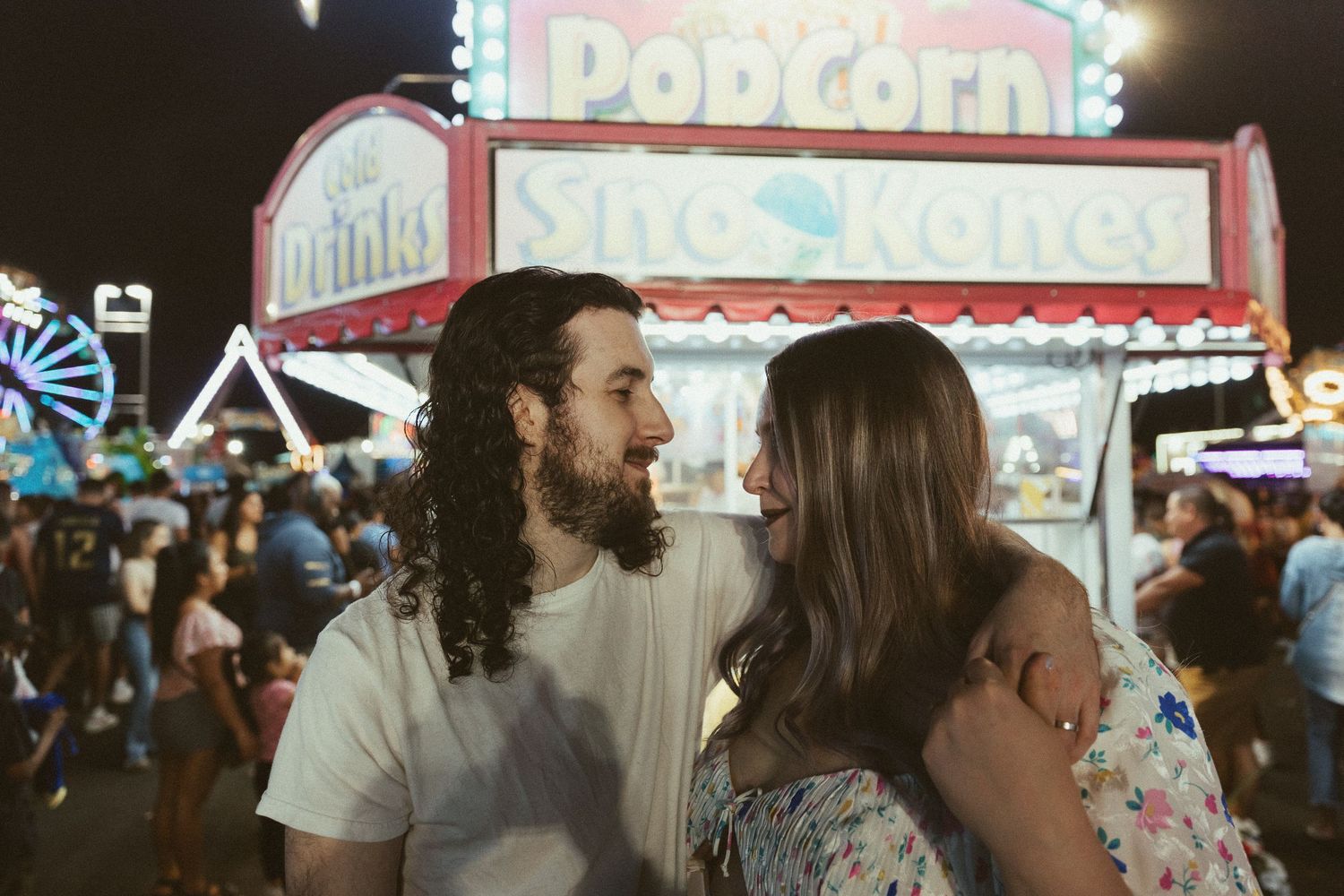 Couple smiling at each other at a colorful night carnival with popcorn and drinks stands in the background.