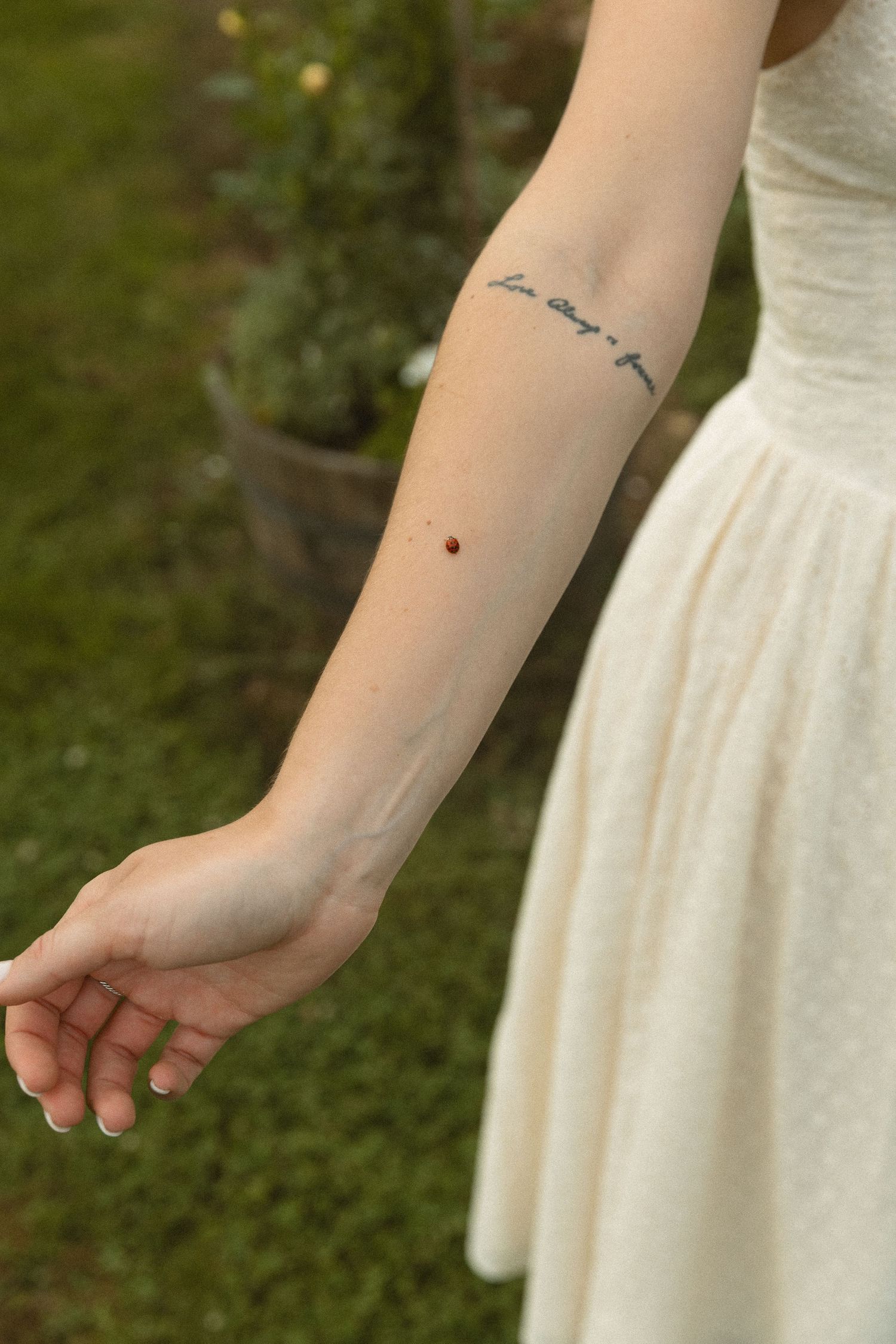 Woman's forearm with cursive script tattoo and small red dot, wearing white dress outdoors.