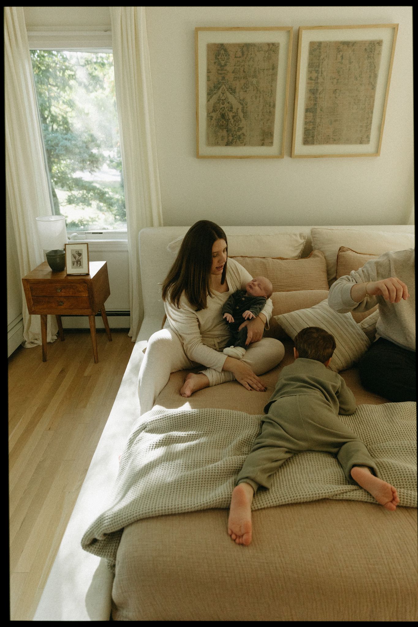 A mother sits cross-legged on a beige sofa holding a newborn while a toddler lies beside her in a bright bedroom.