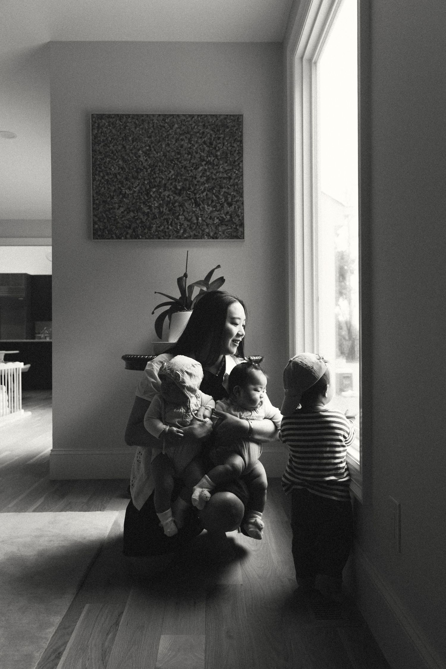 Black and white photo of a smiling mother crouching by a window with three young children in a modern home.