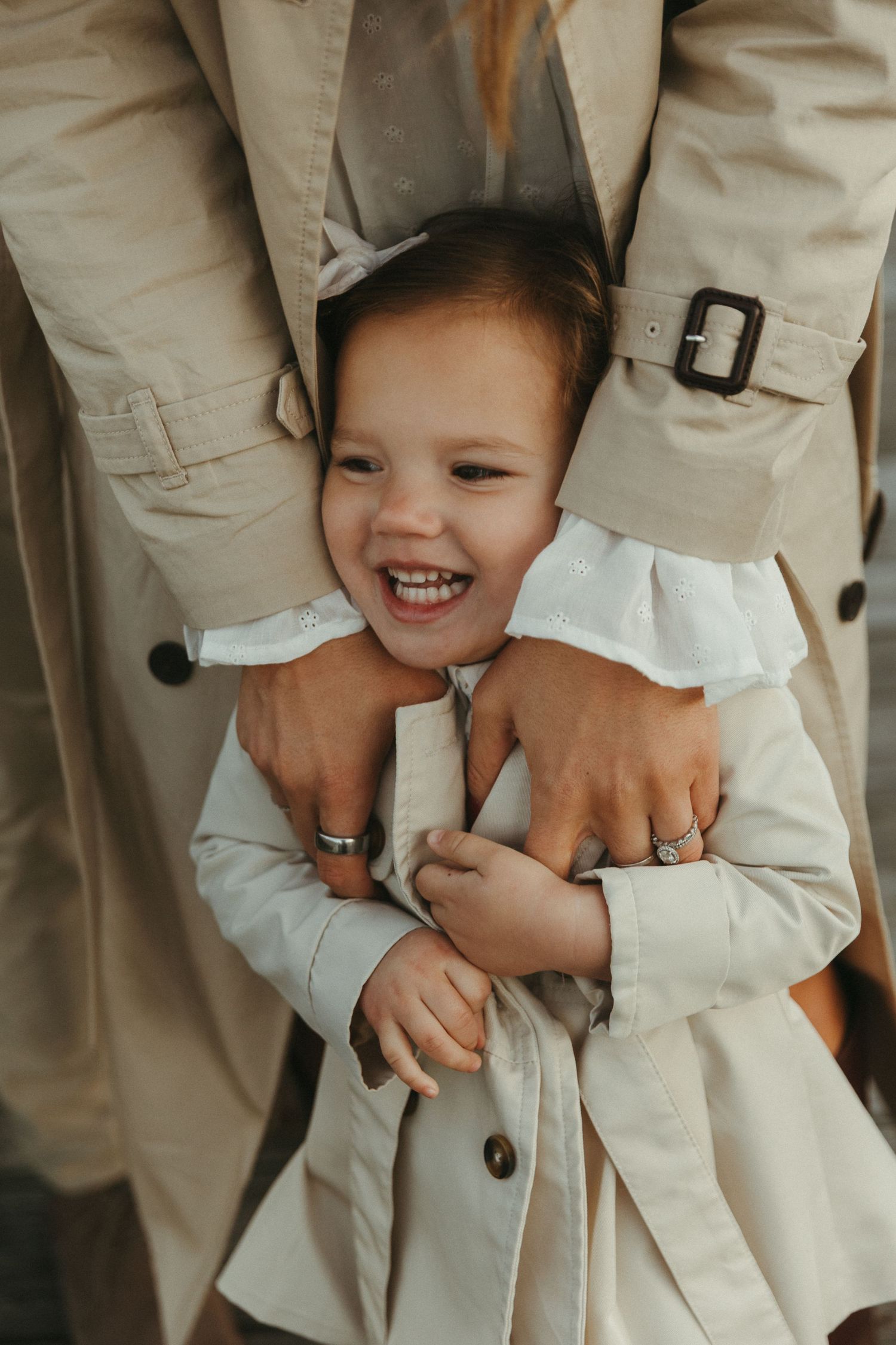 A joyful toddler girl laughs while being hugged from behind by an adult in a beige trench coat.
