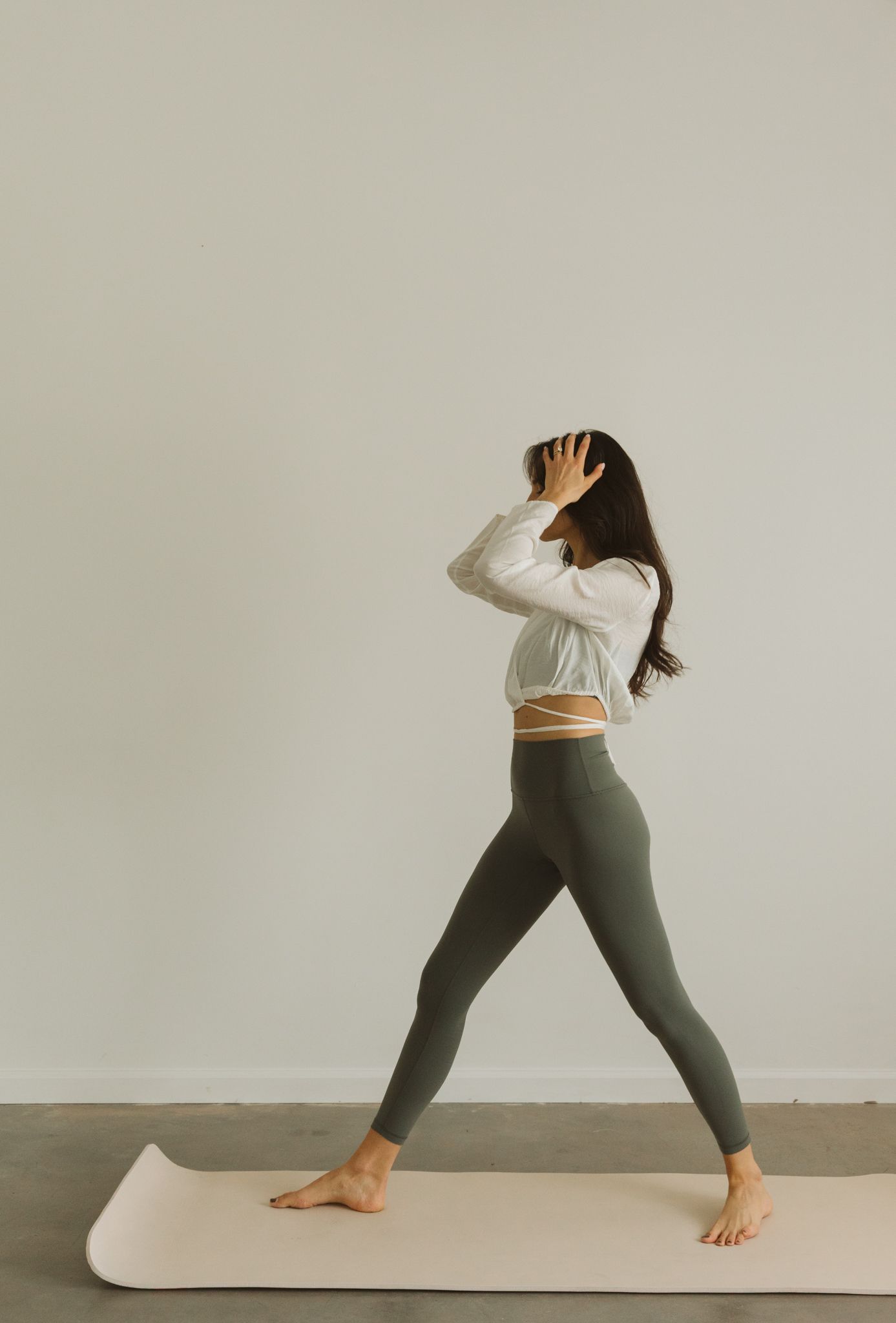 Woman in white crop top and olive leggings practicing yoga on a light pink mat against a neutral wall.