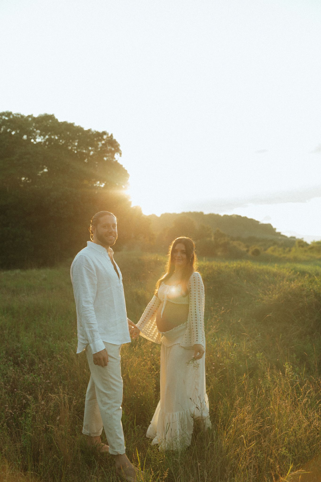 Couple in white outfits holding hands in a sunlit green field at golden hour sunset.