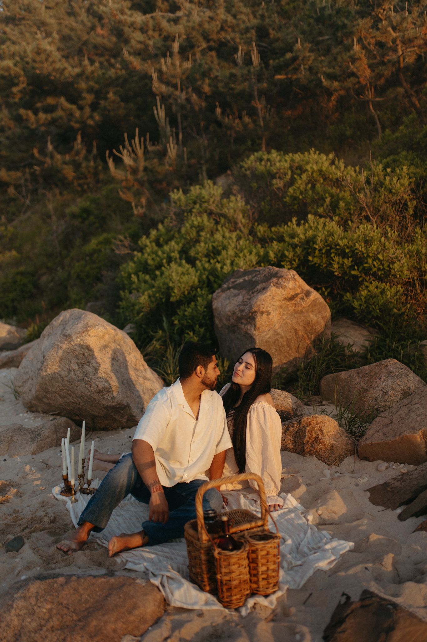Couple sharing a romantic outdoor picnic among boulders and greenery at golden hour sunset.