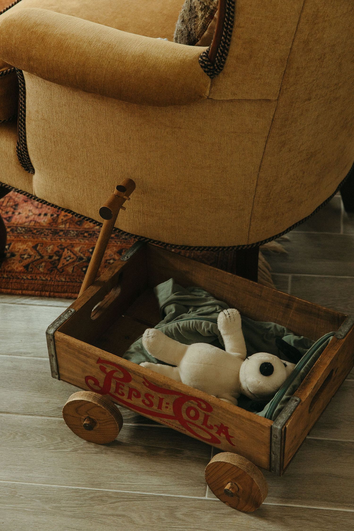 Vintage Pepsi-Cola wooden crate wagon with wheels holding a stuffed bunny toy beside a retro yellow armchair.