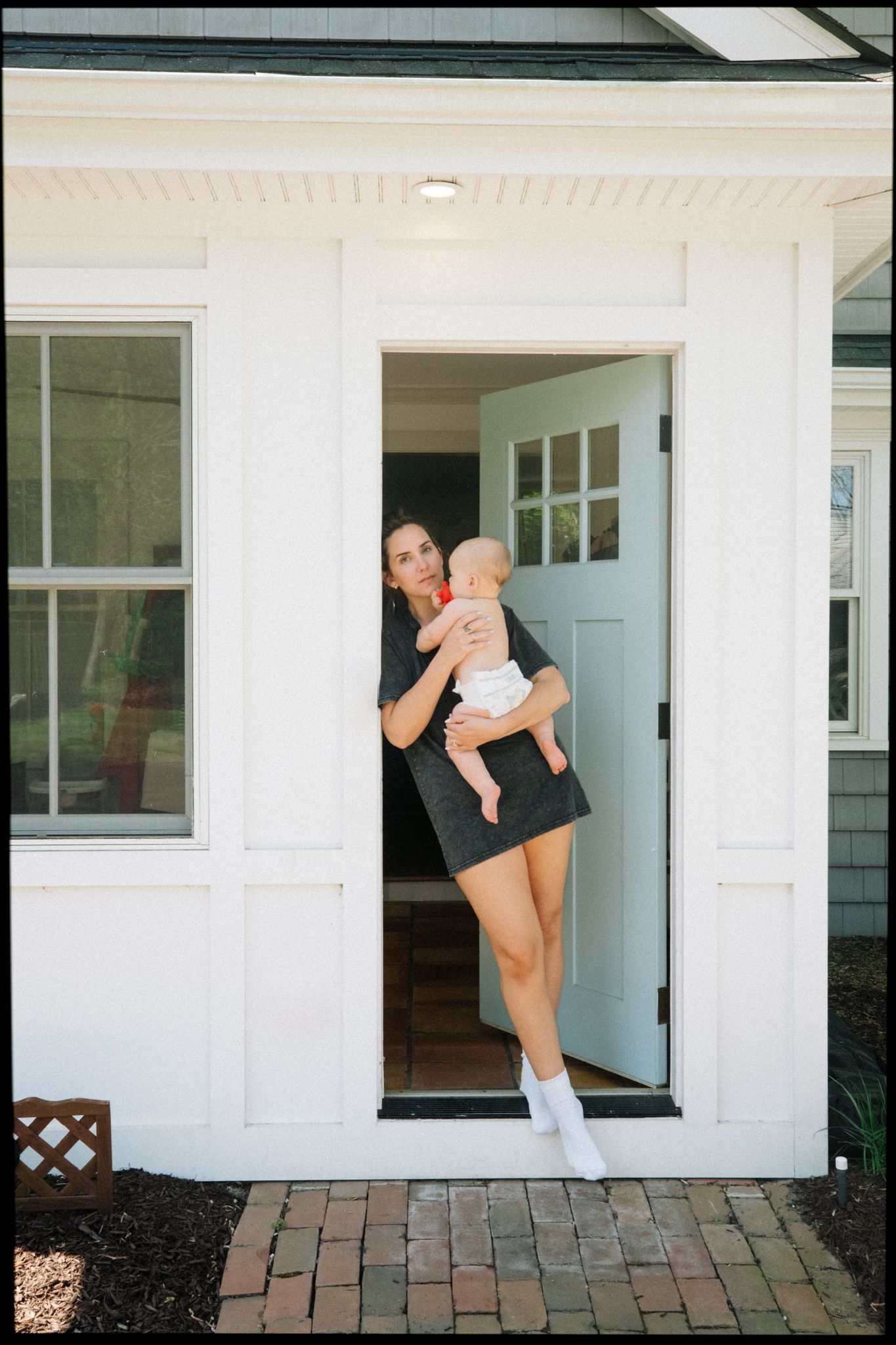 Young mother in black dress holding infant baby while standing in open white cottage door.