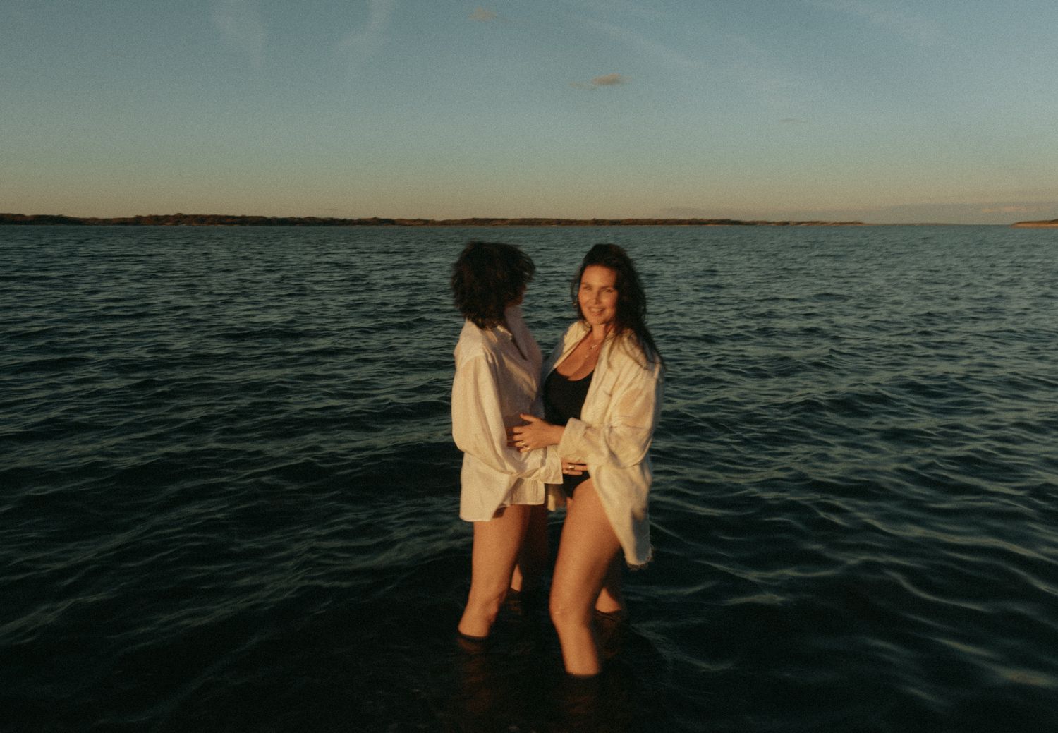 Two women stand together in calm ocean waters at sunset, wearing white shirts and swimwear.