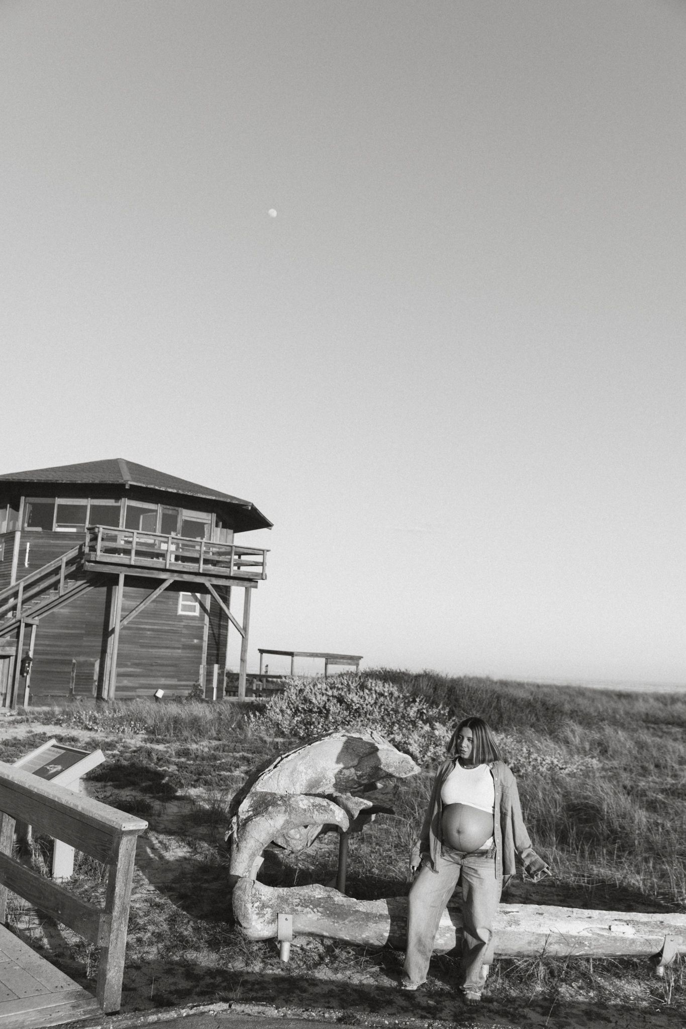 Black and white photo of pregnant woman leaning on driftwood near coastal lifeguard tower at dusk.