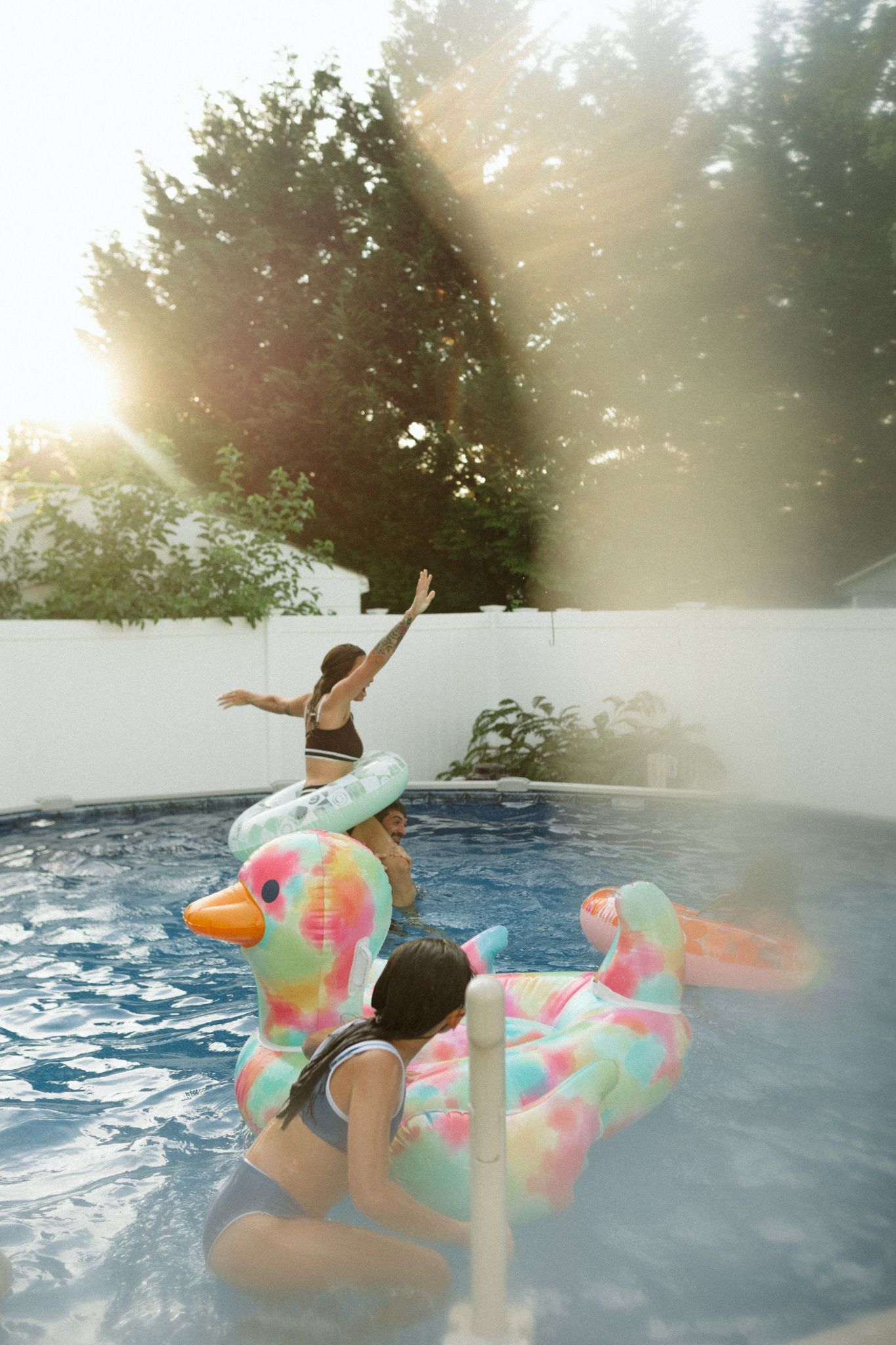 People playing on colorful inflatable flamingo floats in a backyard pool at sunset.