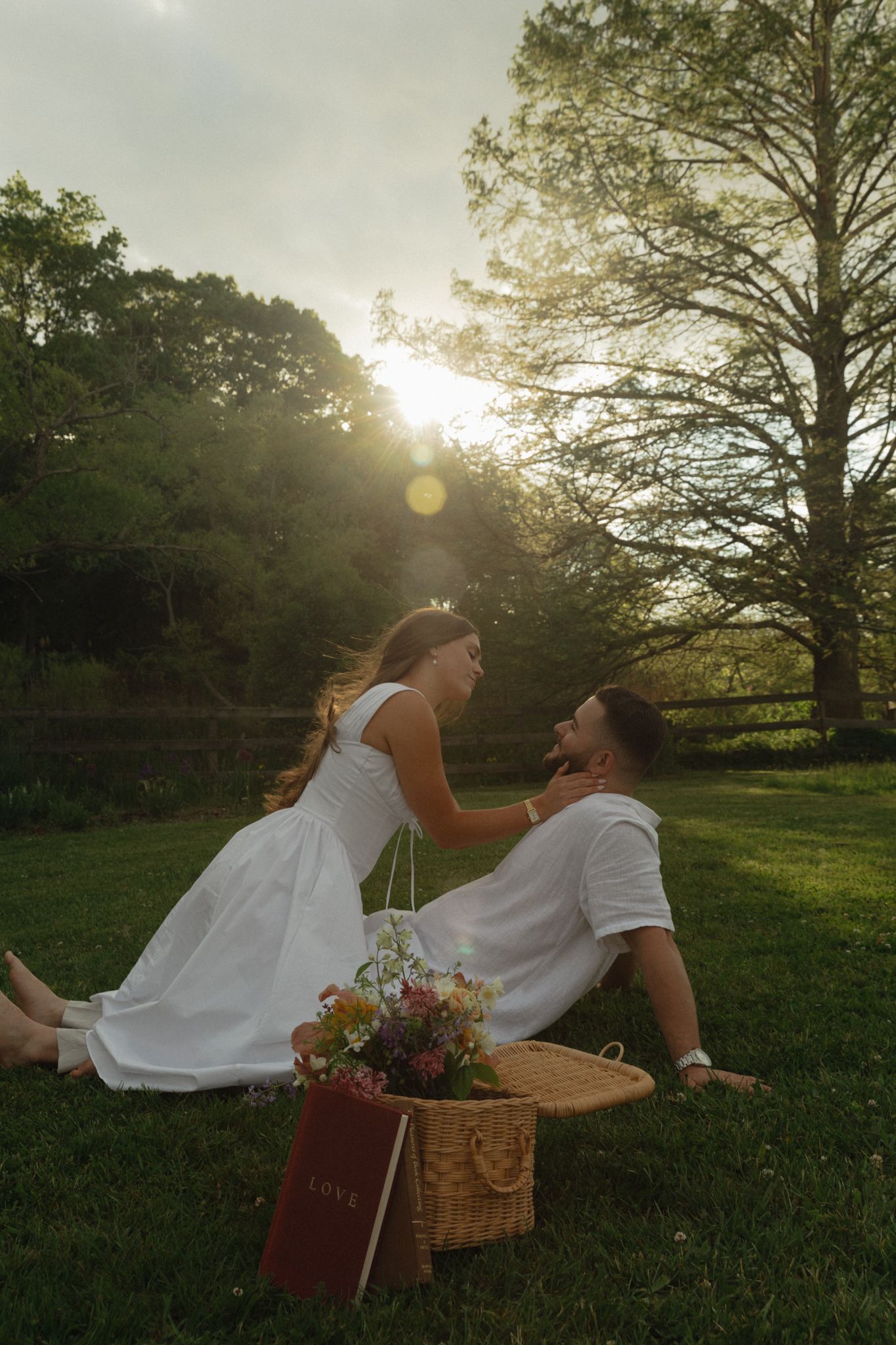 Couple in white outfits sharing a romantic sunset picnic on green grass with a wicker basket and wildflowers.