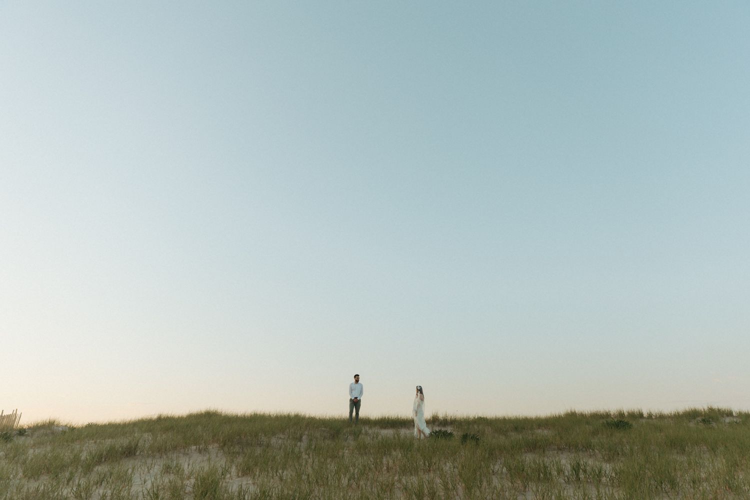 Couple standing on grassy dunes at sunset with a vast clear sky backdrop during an outdoor wedding shoot.