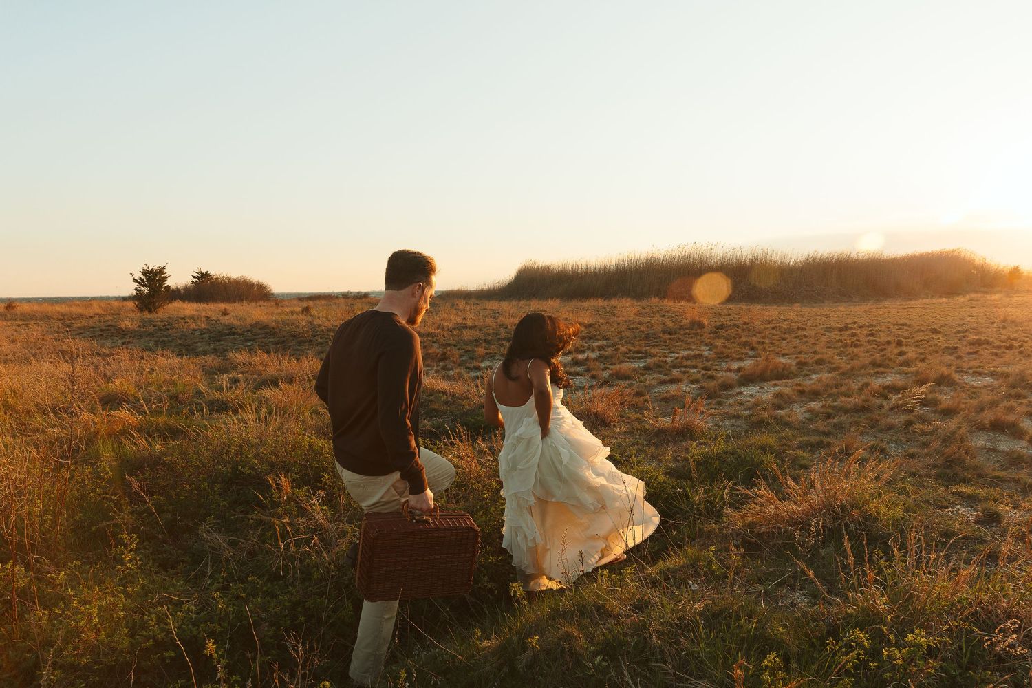 Couple walking hand in hand through golden field at sunset, woman in white dress, man carrying vintage suitcase.