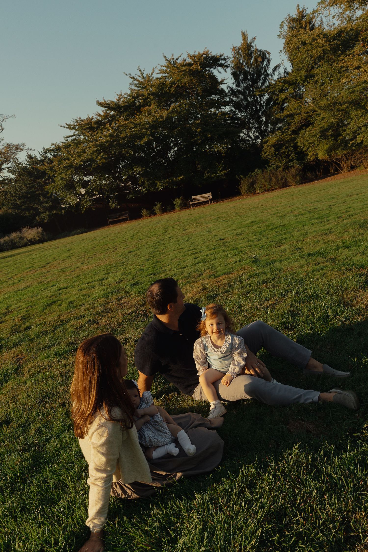 A family relaxes on a sunny green hillside at golden hour, with two children and a parent sitting together.