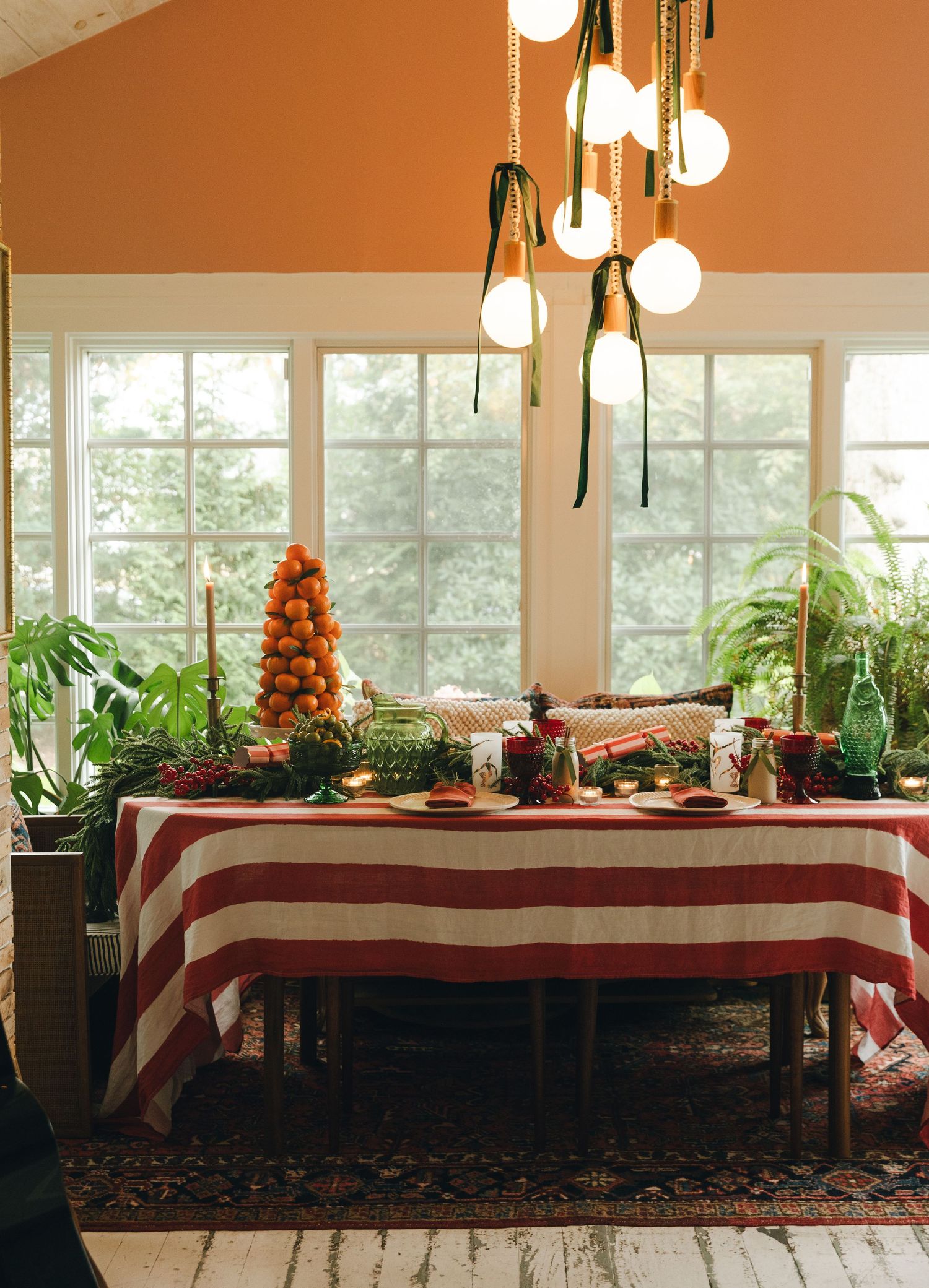 Festive dining table with red and white striped tablecloth, citrus topiary, candles, and globe pendant lights in a sunlit room.
