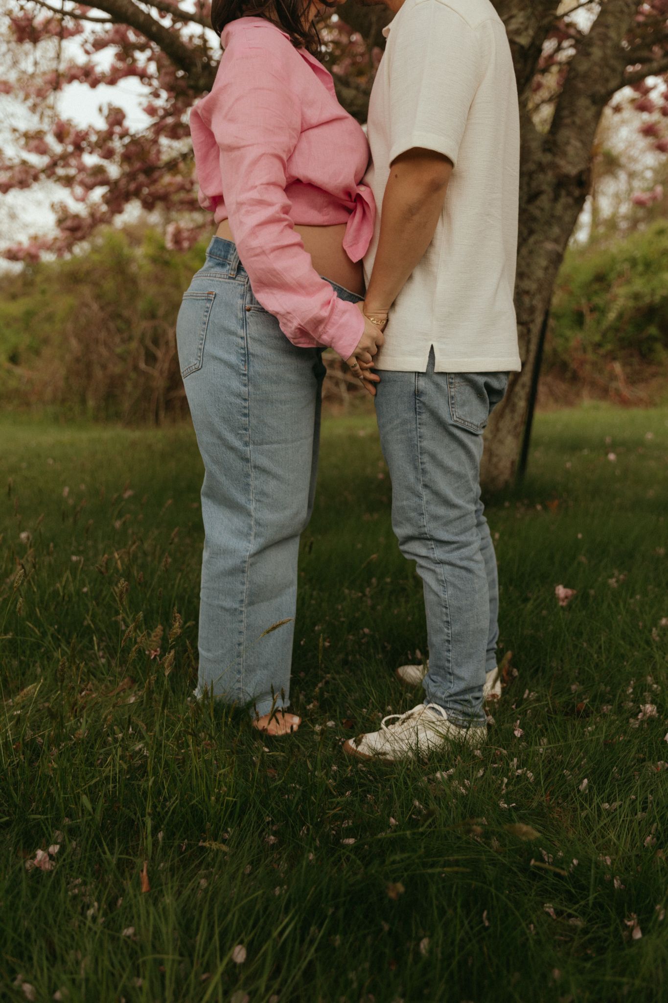 Lower body shot of two people standing close together on grass under spring trees.
