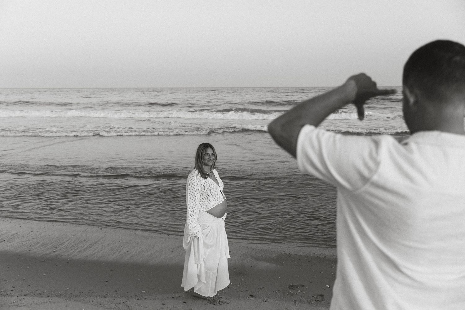 Black and white photo of pregnant woman in white dress posing on beach while partner photographs her.