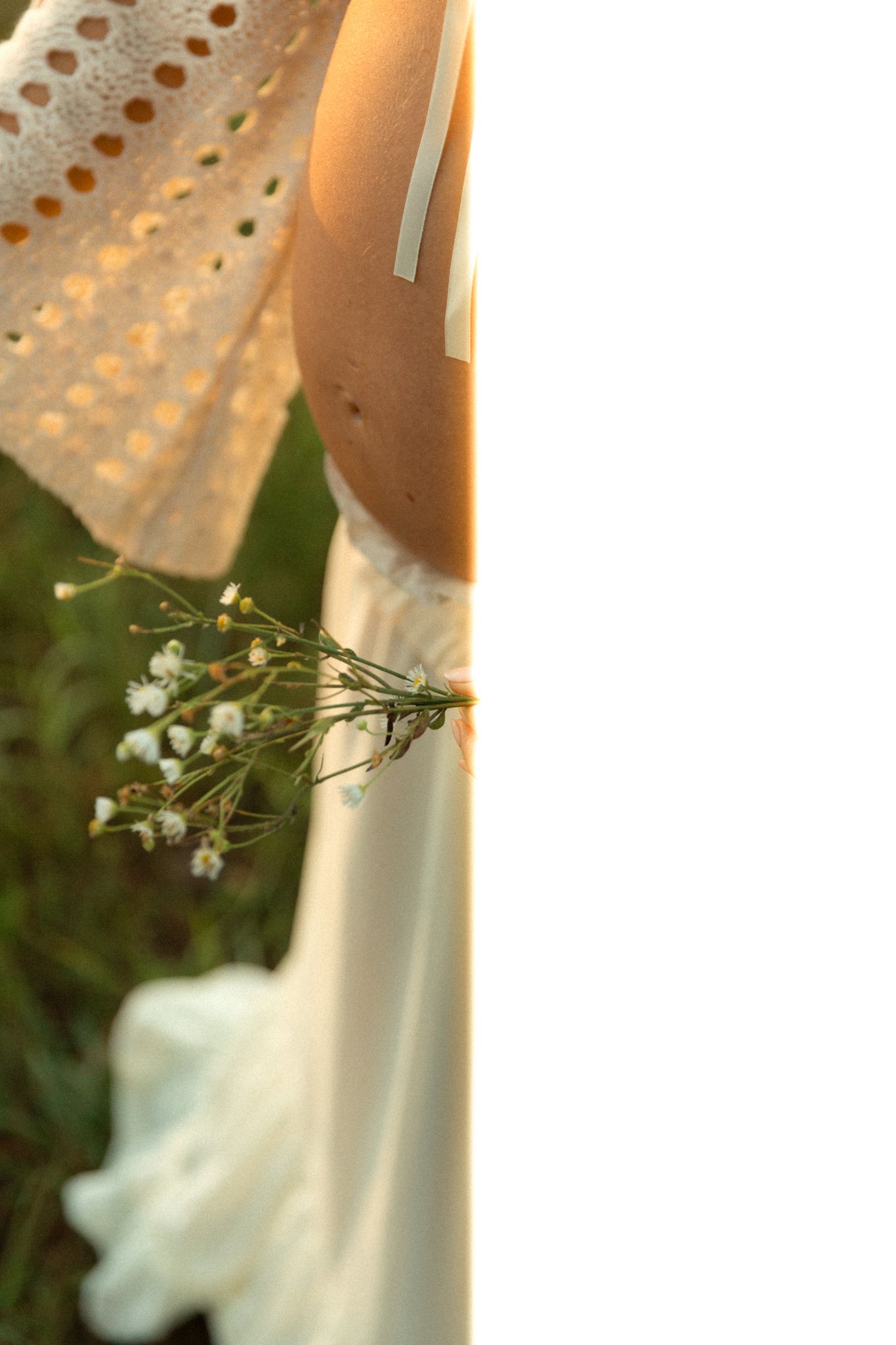 Close-up of pregnant belly with wildflowers held by woman in white lace dress outdoors.