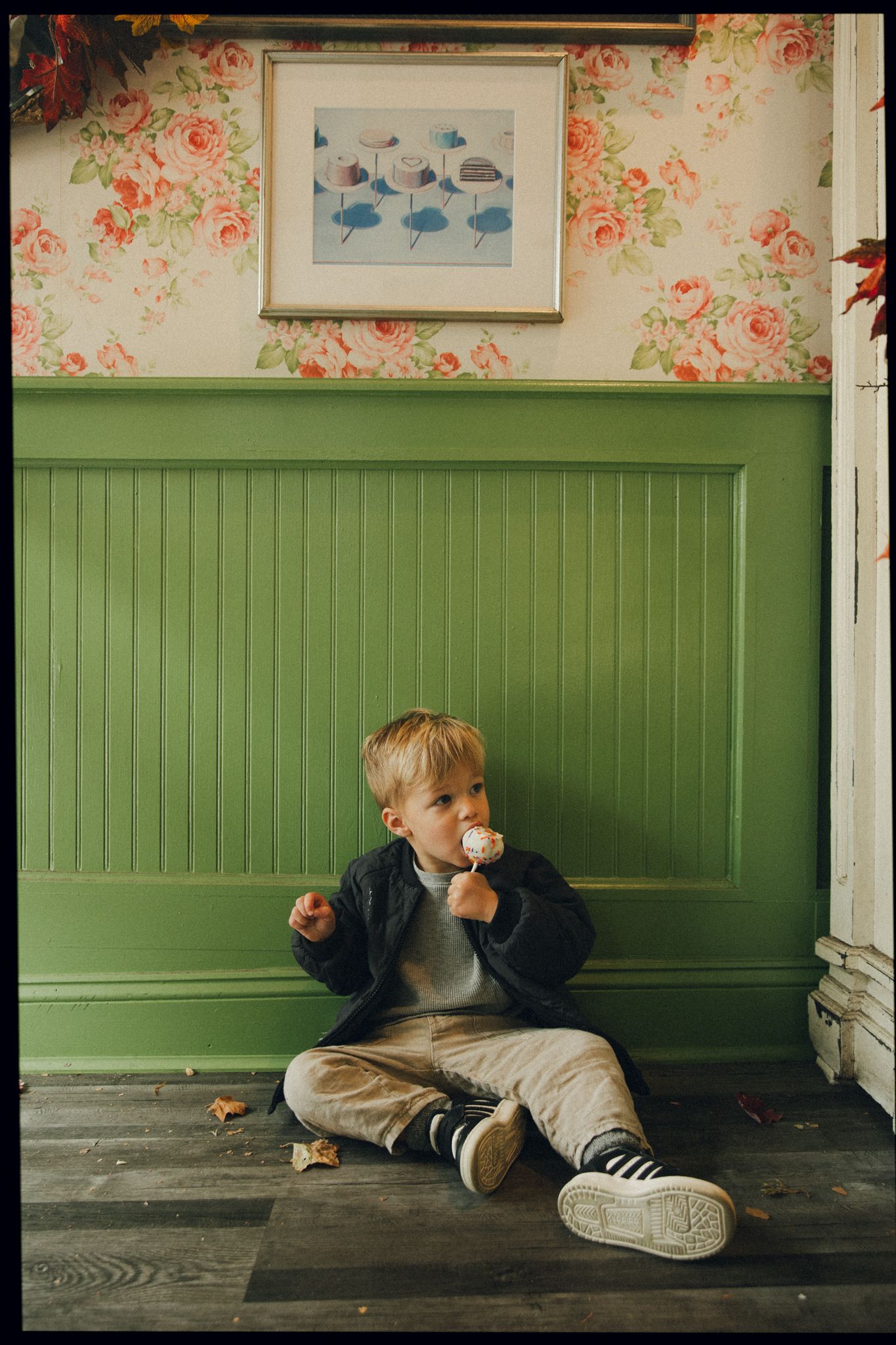 Young toddler boy sitting on wooden floor eating a lollipop against green wainscoting and floral wallpaper.
