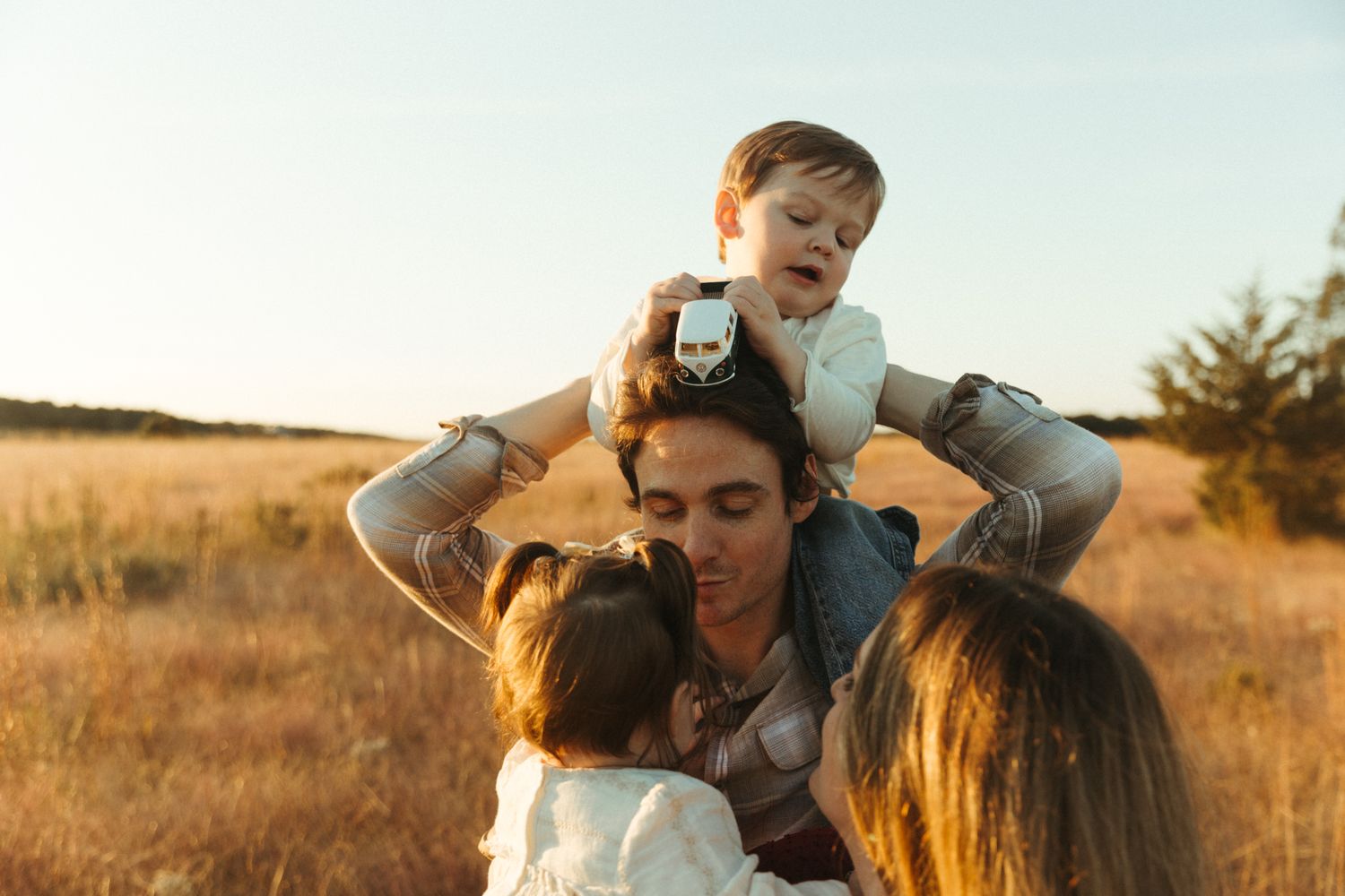Family enjoying golden hour in a field, father with toddler on shoulders embracing children outdoors.