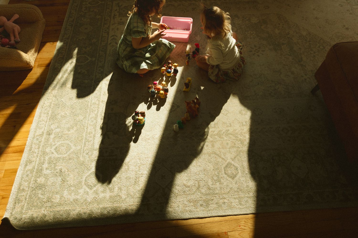 Two children playing with colorful toys on a rug, viewed from above with dramatic sunlight shadows on hardwood floor.