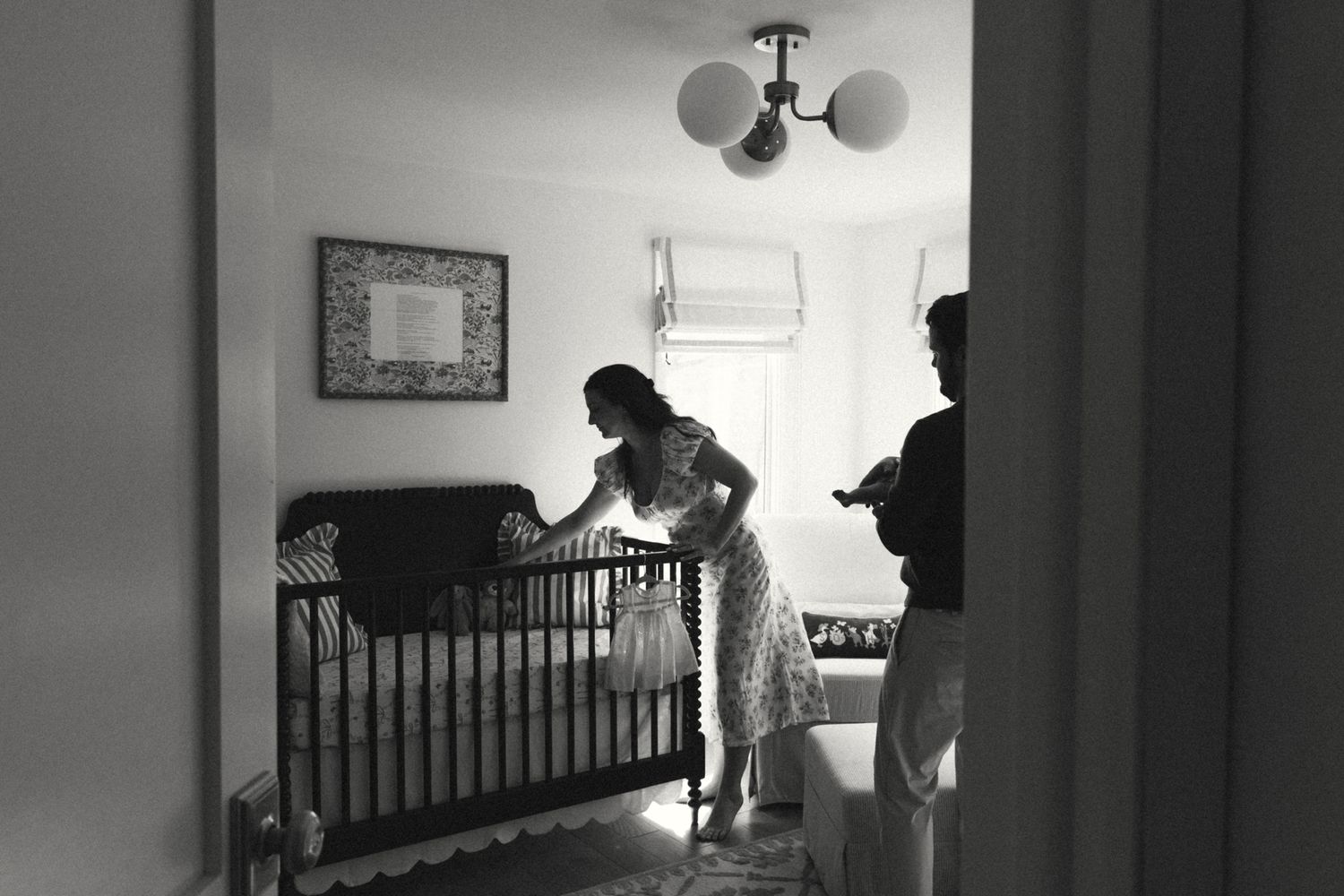 Black and white photo of a woman leaning over a baby crib in a nursery room with soft lighting.