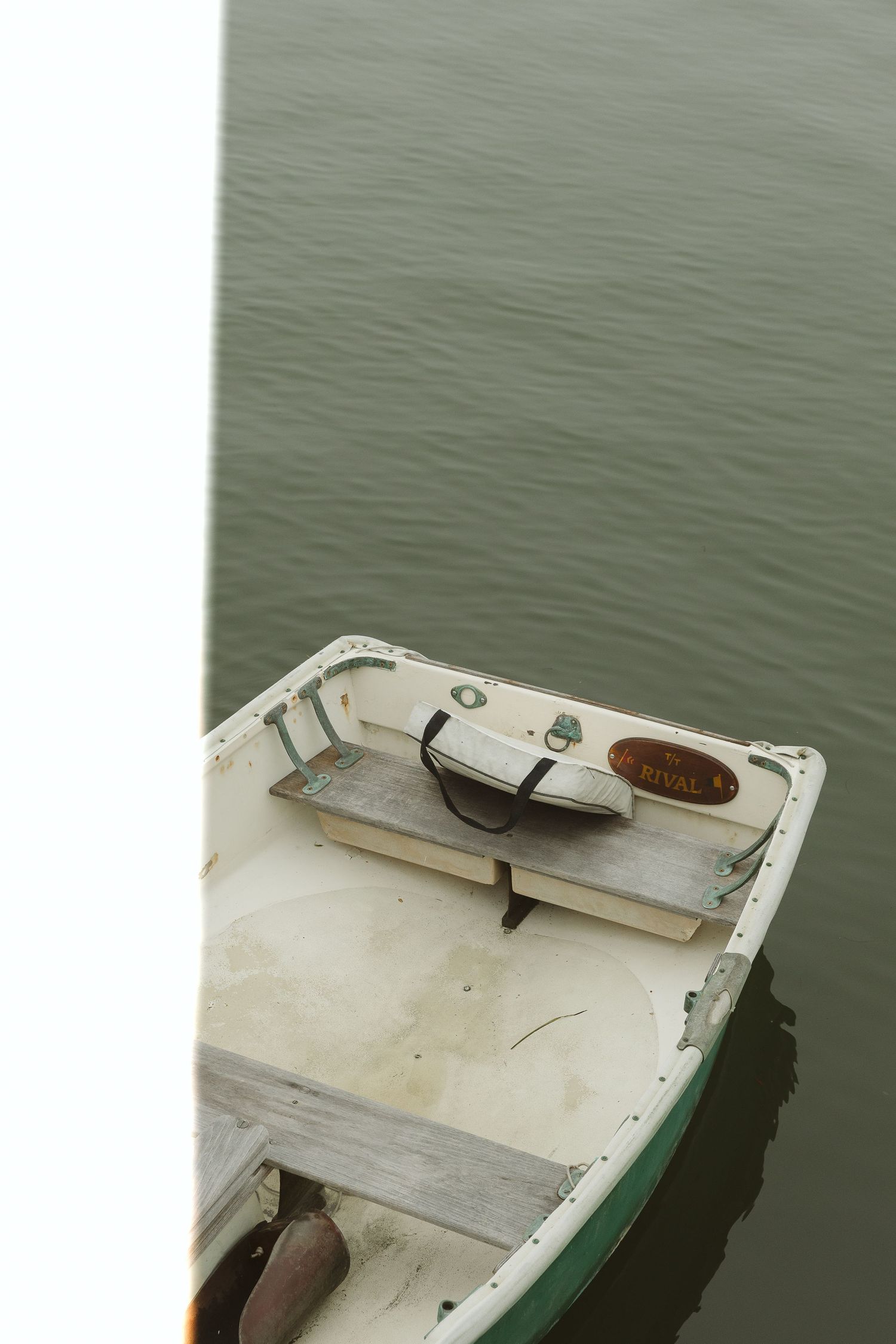 A weathered white rowboat with a wooden seat floats on calm green water near a dock.