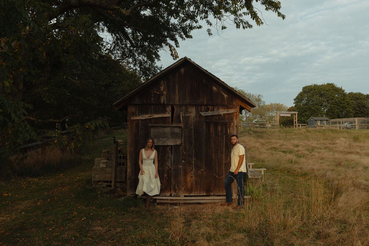 Couple poses by rustic wooden shed in golden field during outdoor farm wedding photography session.