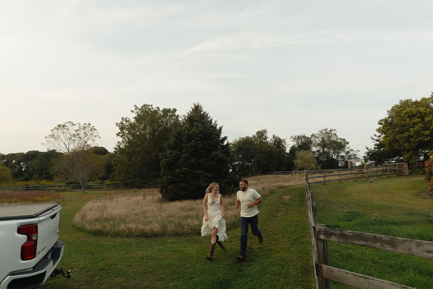 Couple walking hand in hand through a scenic farm with green fields, wooden fence, and trees under a cloudy sky.