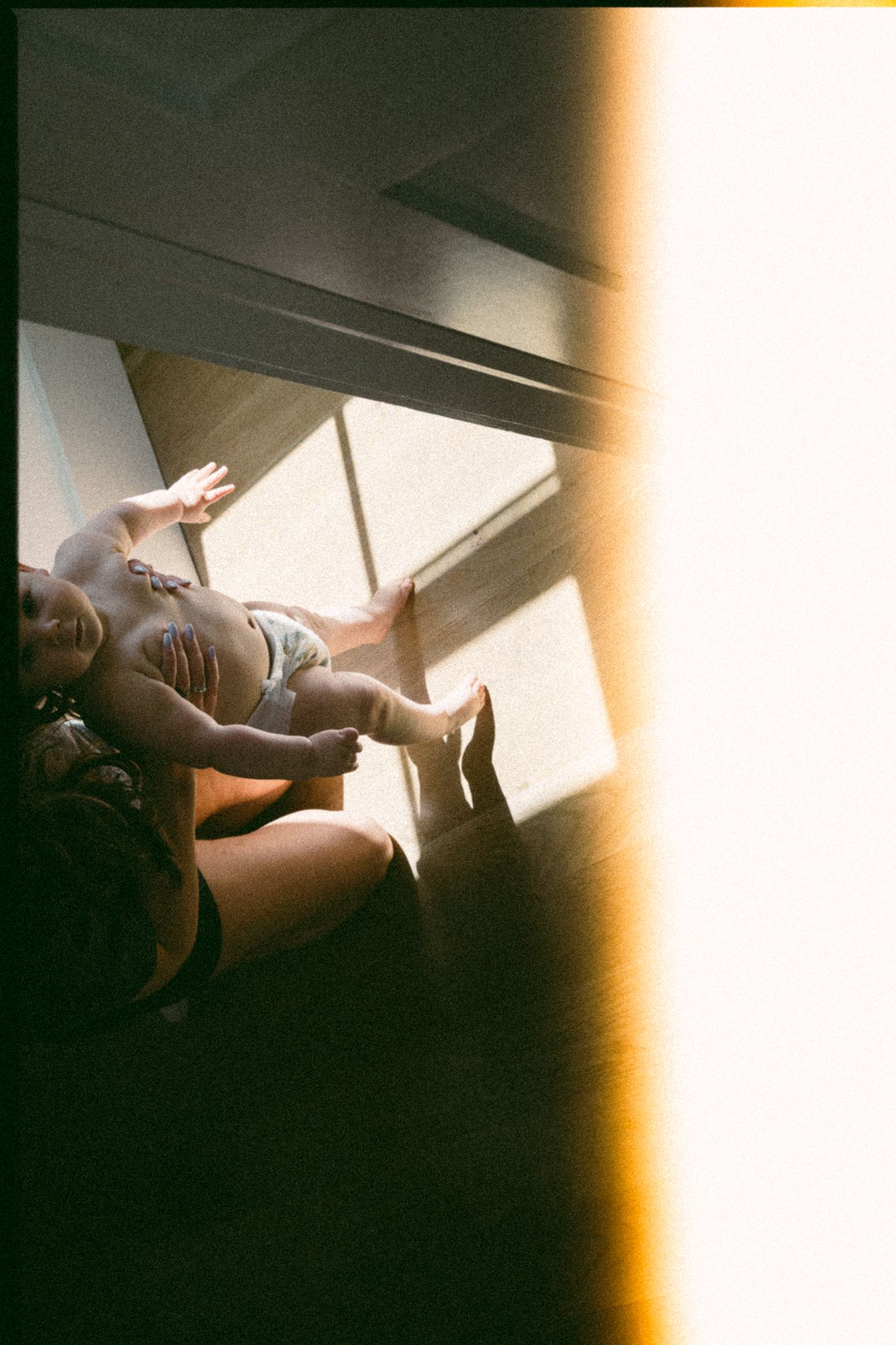 Mother holding baby near sunlit window, captured in warm film photography with artistic light leak effect.