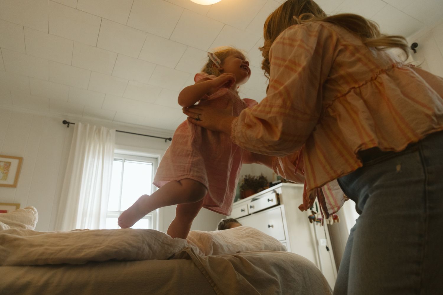 Woman helping young toddler girl stand on a bed in a bright bedroom with white walls and natural light.