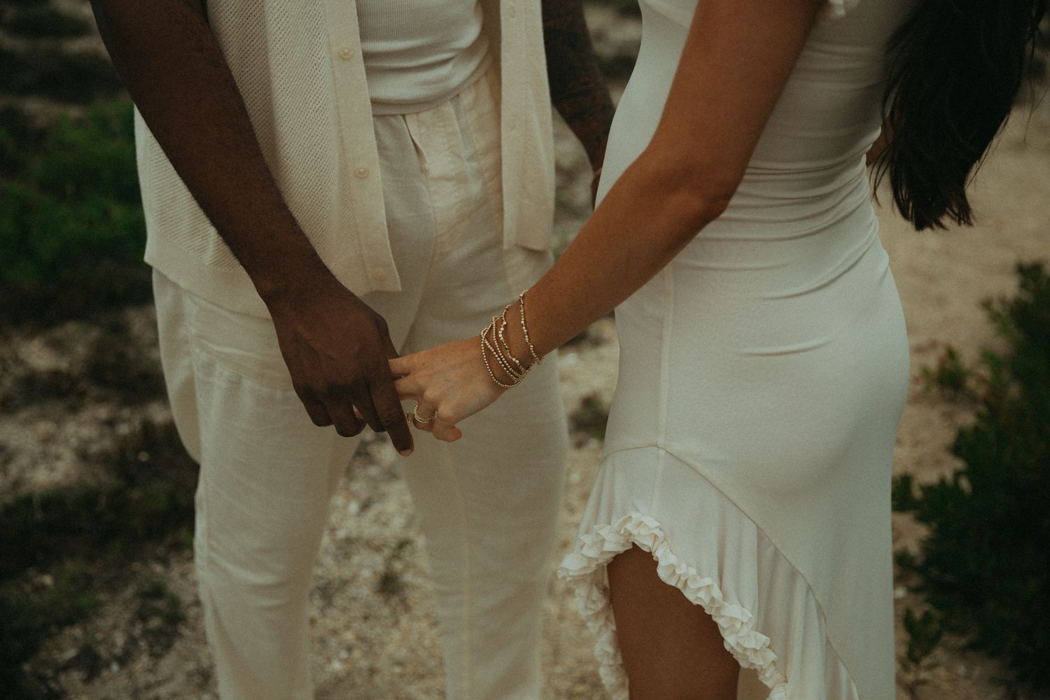 Interracial couple holding hands in matching white outfits, woman wearing gold bracelets, outdoor setting.
