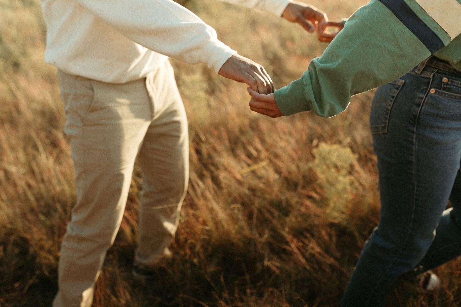 Couple holding hands while walking through golden dry grass fields during a warm sunset outdoor photoshoot.