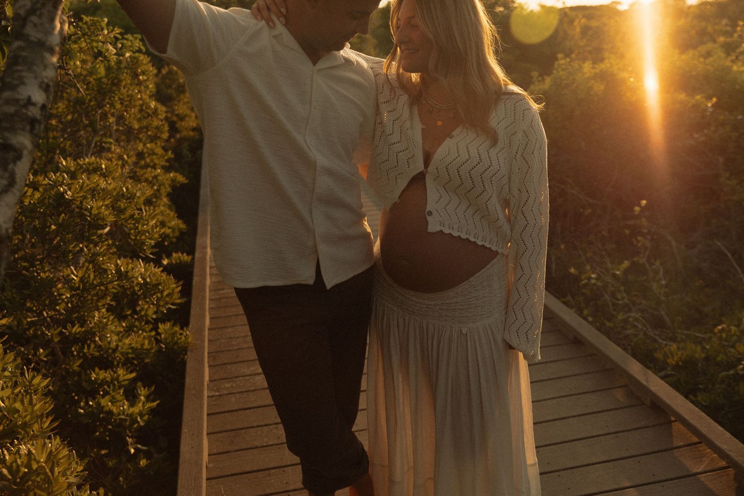 Couple posing on a wooden bridge at golden sunset, woman in white lace maternity dress.