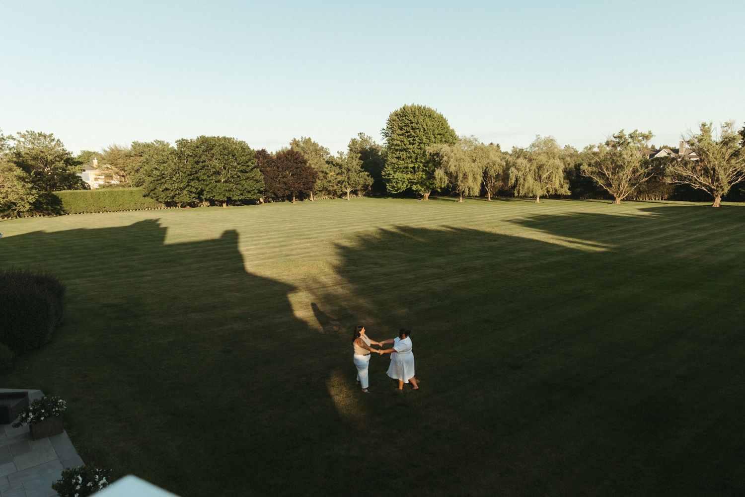 Aerial drone view of couple embracing on vast green lawn casting long shadows at sunset.