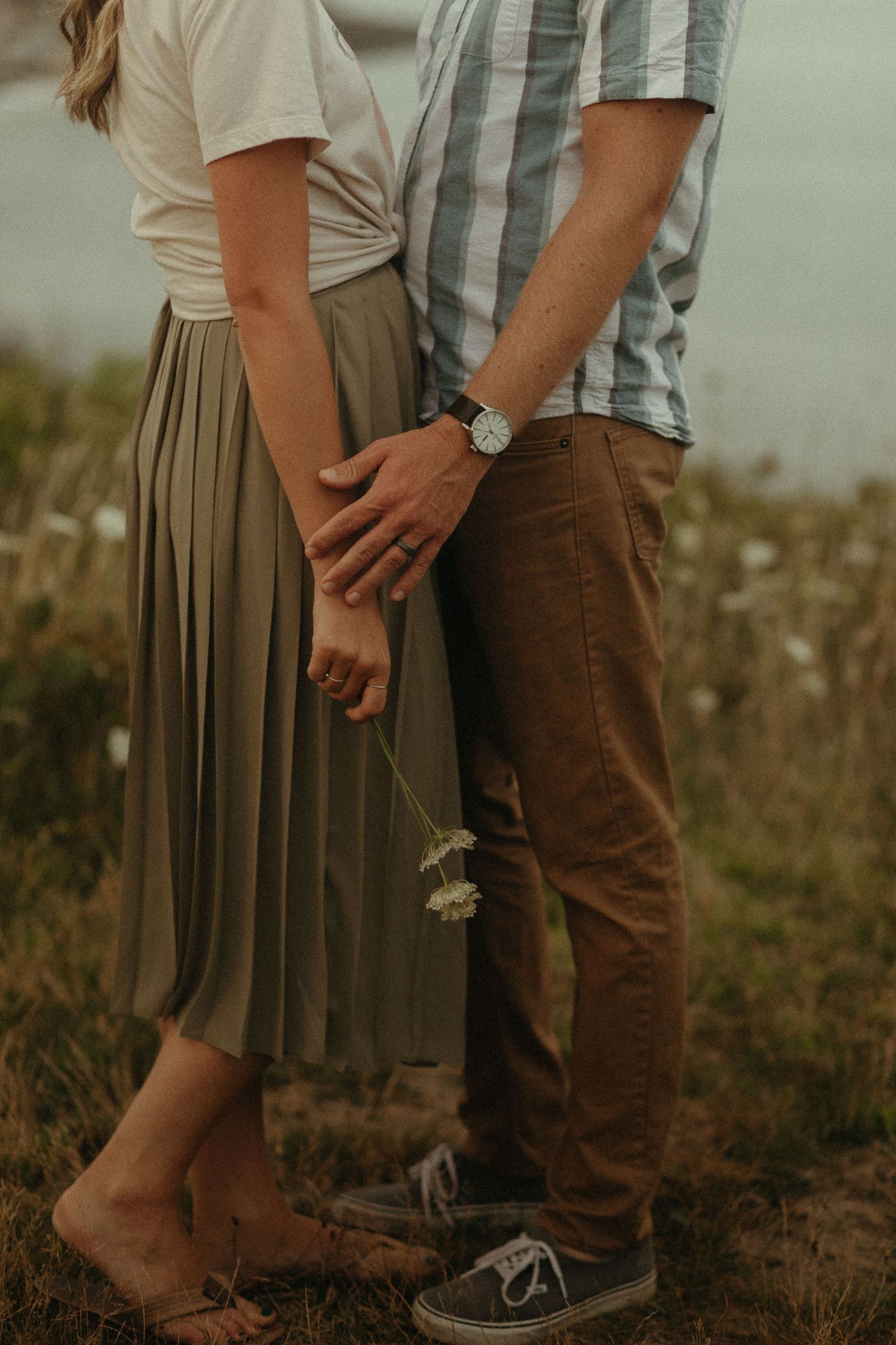 Romantic couple holding hands in a meadow, woman in olive skirt holding a small wildflower.